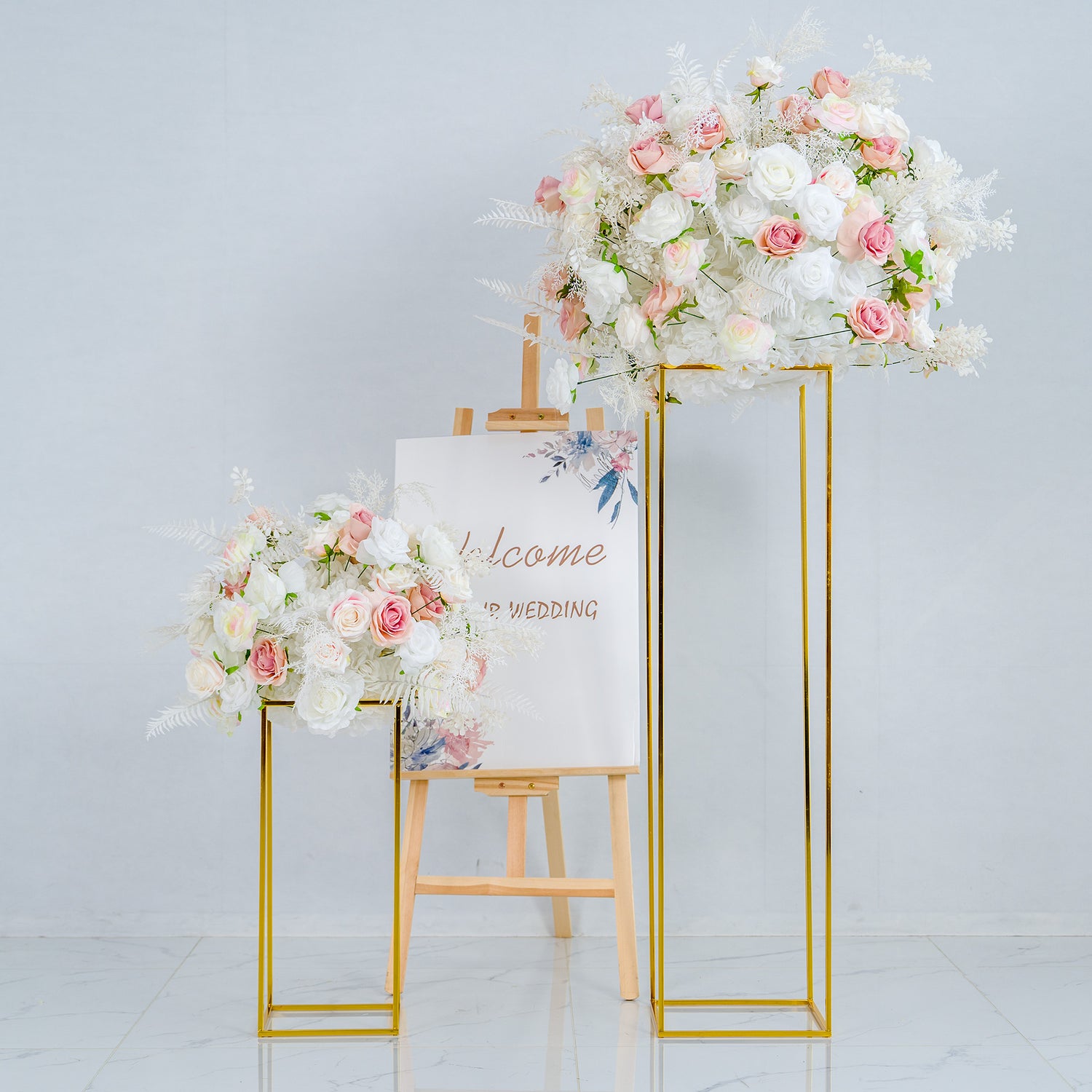 This photo presents a charming wedding - themed decor setup. Two golden - framed stands hold lush floral arrangements composed of white and pink roses, along with white foliage. Between them is an easel with a sign that reads "Welcome to our wedding," adorned with delicate floral illustrations. The overall style is elegant and romantic, perfect for a wedding entrance.