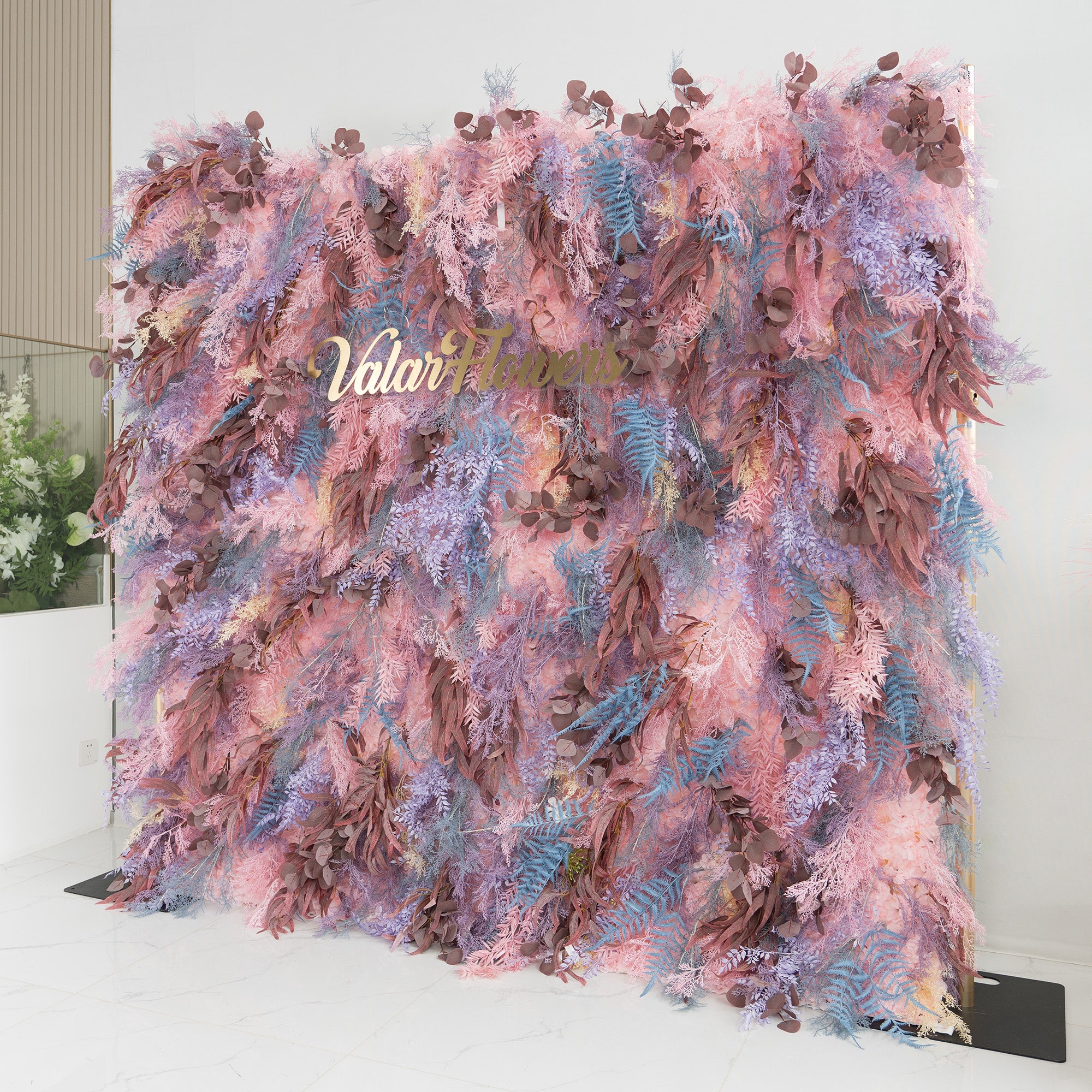 This backdrop is a lush, colorful creation, densely packed with an array of foliage in soft pinks, purples, blues, and earthy browns. The elegant "Valar Flowers" script in gold lettering is prominently displayed in the center. Ideal for events like weddings or upscale parties, it exudes a romantic and dreamy style, adding a touch of enchantment to any setting.