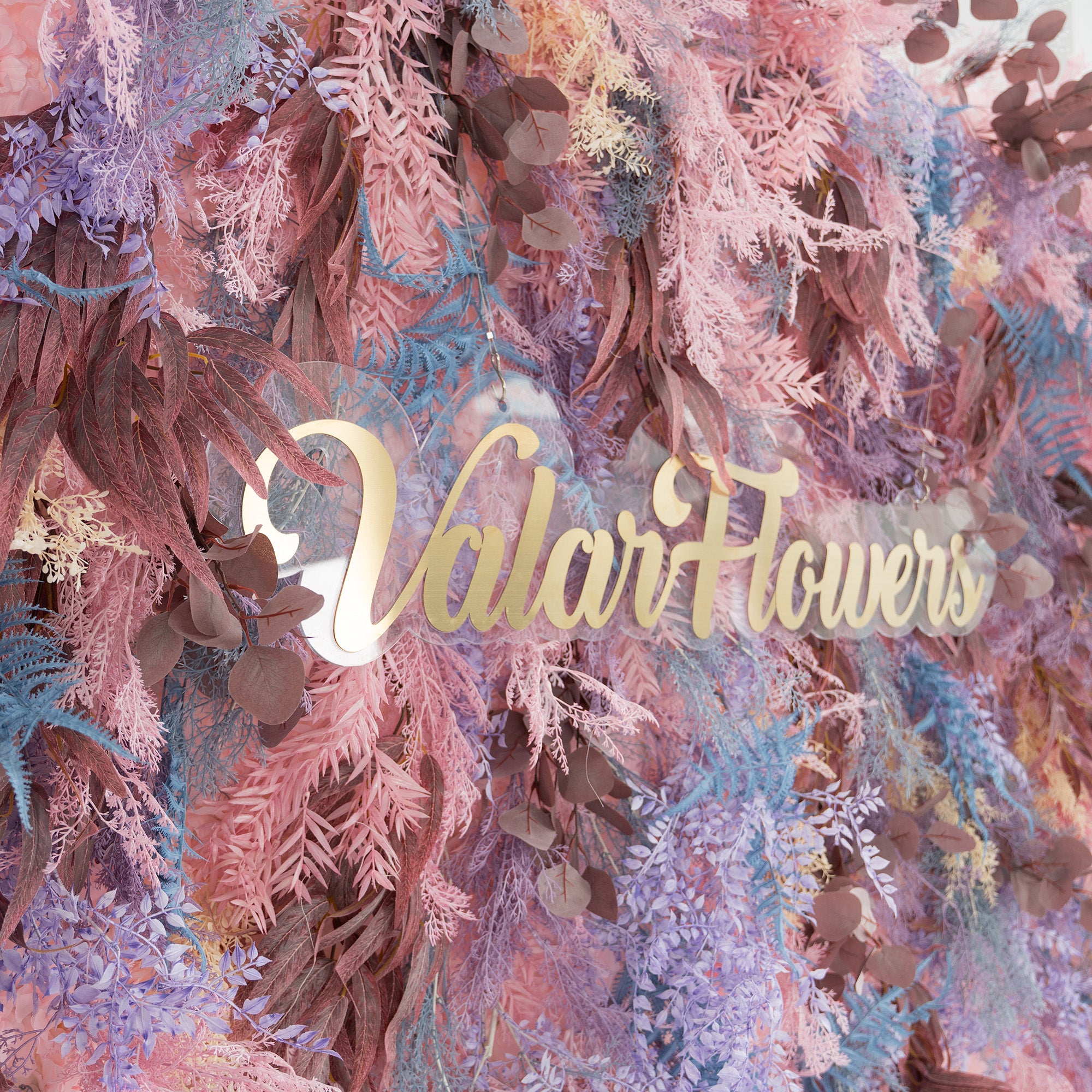 This photo showcases a lush, vibrant foliage backdrop. It features a mix of fern - like fronds in shades of blue and purple, along with eucalyptus leaves in earthy brown tones, all blended with soft pink foliage. The elegant gold text "Valar Flowers" stands out prominently. Ideal for weddings, parties, or upscale events, it exudes a romantic and whimsical style.