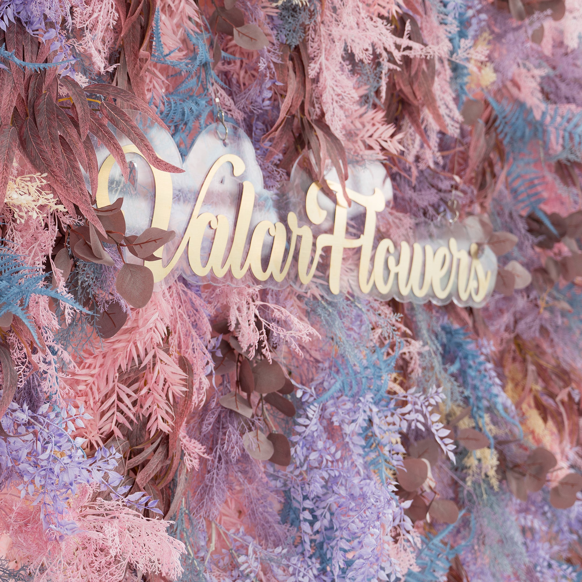 This photo showcases a lush, colorful foliage backdrop. It features a mix of fern - like fronds in shades of blue and purple, along with eucalyptus leaves in earthy brown tones, all blended with soft pink foliage. The elegant gold text "Valar Flowers" stands out prominently. Ideal for weddings, parties, or upscale events, it exudes a romantic and whimsical style.