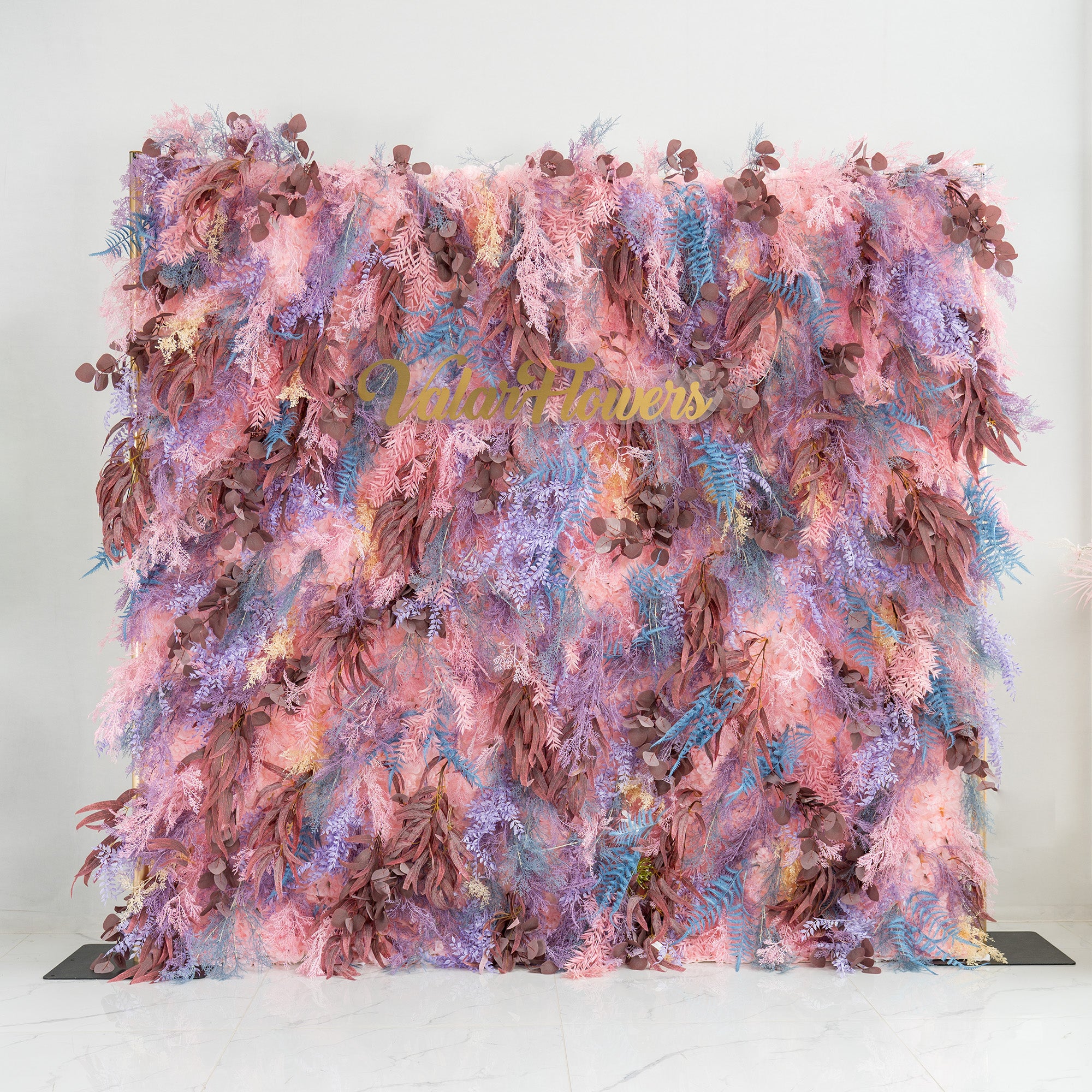 This photo showcases a lush foliage backdrop, a vibrant blend of plants. It features fern - like fronds in blue and purple, alongside eucalyptus - like leaves in earthy browns, all intertwined with soft pink foliage. The words "Valar Flowers" in elegant gold lettering stand out. Ideal for weddings, parties, or upscale events, it exudes a romantic and whimsical style.