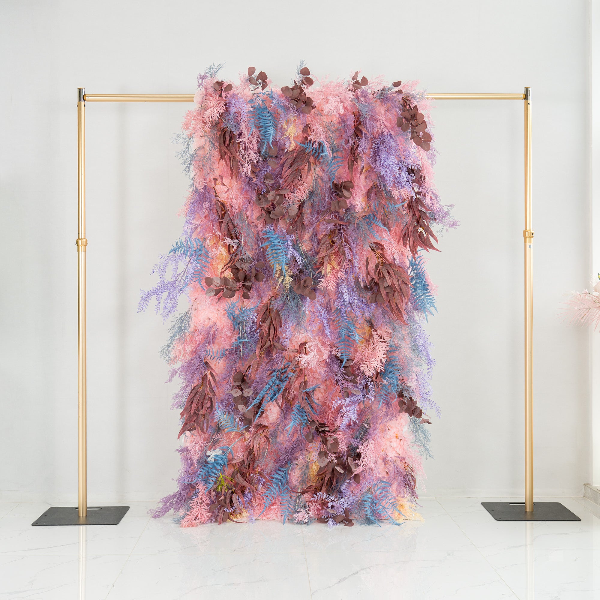 This photo features a lush foliage backdrop supported by a golden frame. It combines fern - like fronds in shades of blue and purple with eucalyptus - like leaves in earthy brown tones, all interwoven with soft pink foliage. The pastel color palette creates a dreamy look. Ideal for weddings, parties, or upscale events, it exudes a romantic and whimsical style.