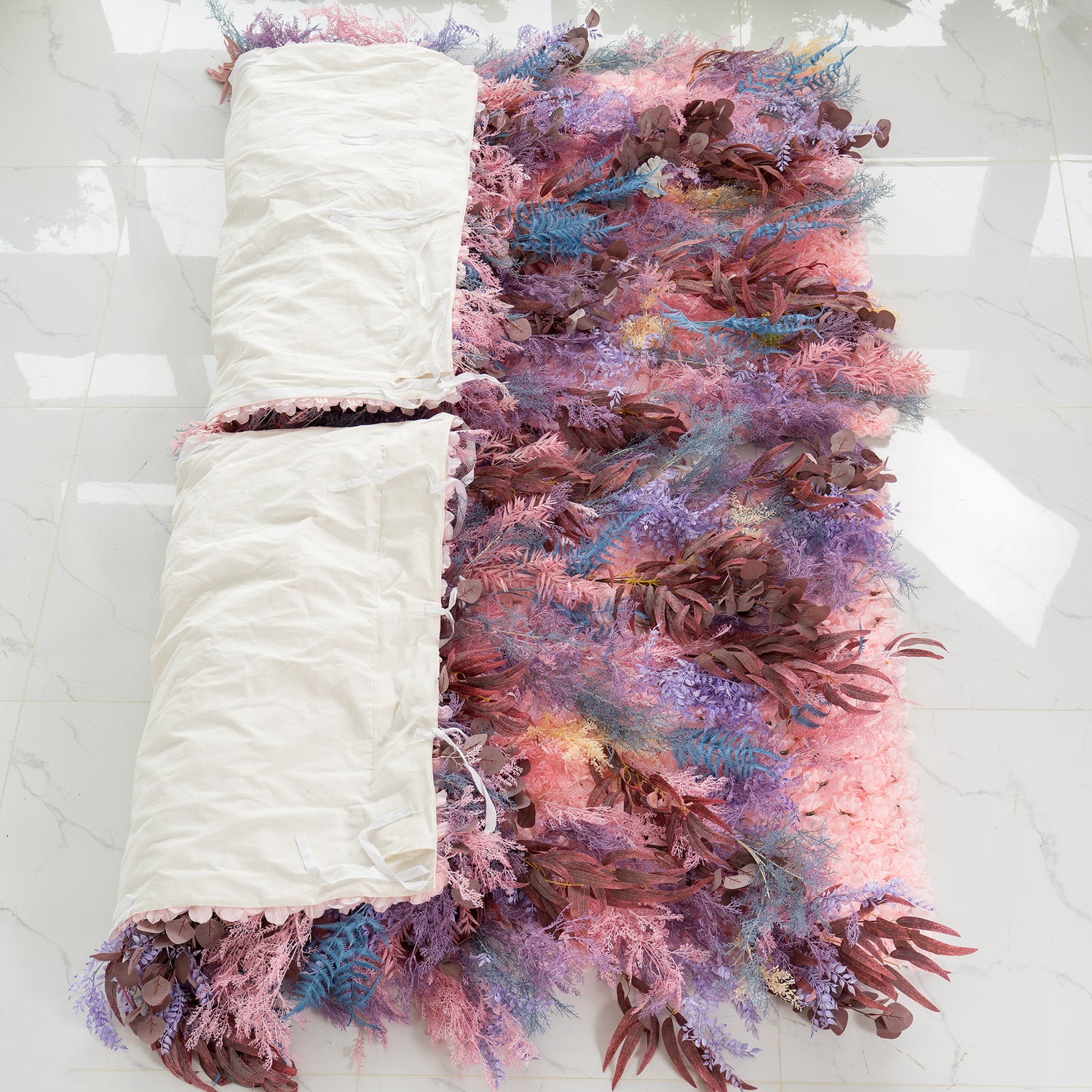 This photo shows a foliage backdrop partially rolled up in white fabric. The backdrop features a rich mix of plants, including fern - like fronds in shades of blue and purple, along with eucalyptus - like leaves in earthy browns and soft pink foliage. Its pastel color palette gives it a dreamy feel. Ideal for events like weddings or parties, it exudes a romantic and whimsical style.