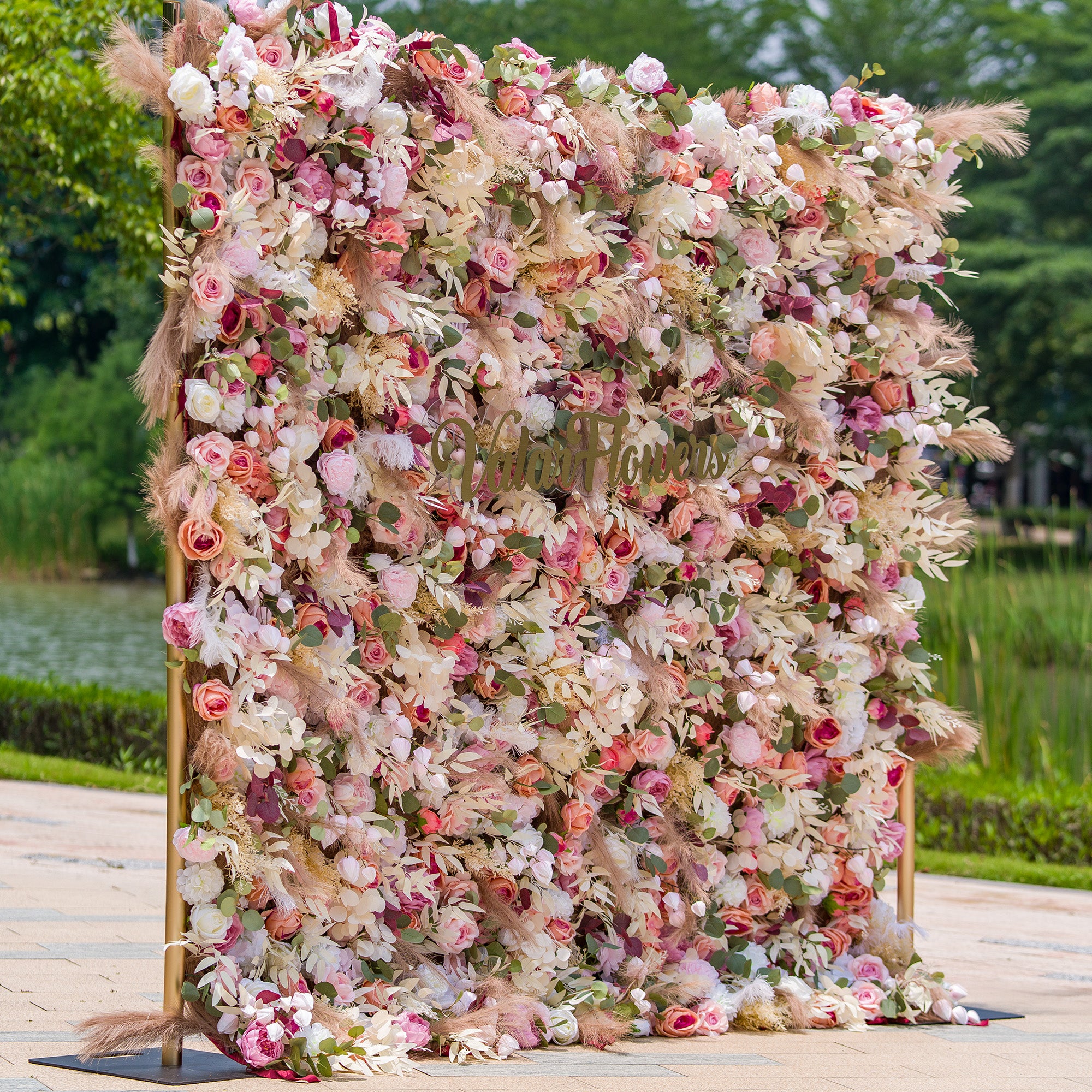 This photo showcases a large, lush floral backdrop. It is densely filled with an assortment of roses, peonies, and other blooms in soft pinks, whites, and hints of peach, interspersed with green foliage and pampas grass. The sign “ValarFlowers” is prominently displayed. Ideal for weddings, garden parties, or upscale events, it creates a dreamy, romantic atmosphere.