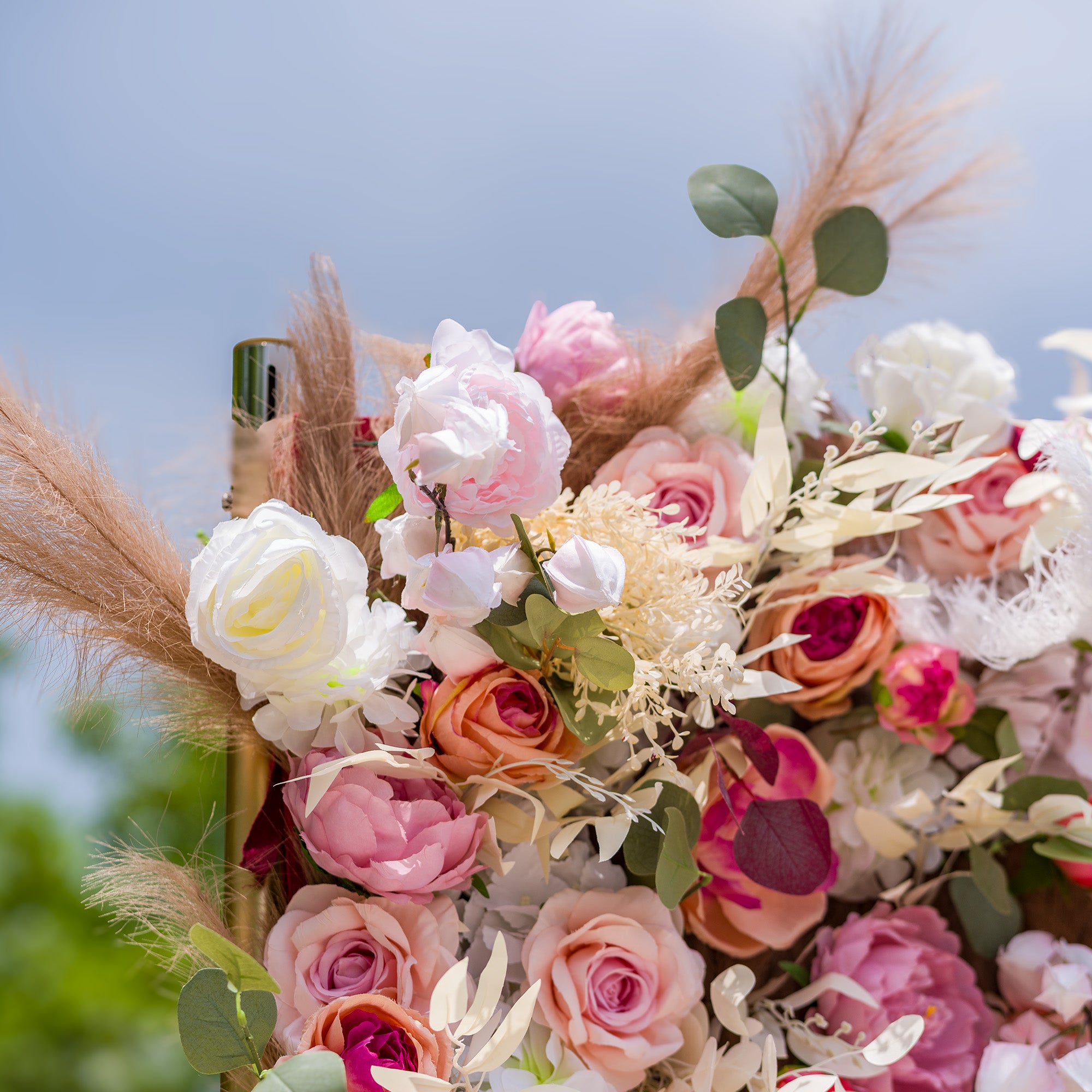 This photo showcases a lush arrangement of artificial flowers in soft hues of pink, white, and peach, including roses and peonies. Interspersed with green eucalyptus leaves and fluffy pampas grass, it exudes a romantic and elegant vibe. Ideal for weddings, bridal showers, or upscale garden parties, it can serve as a beautiful backdrop or centerpiece to create a dreamy atmosphere.