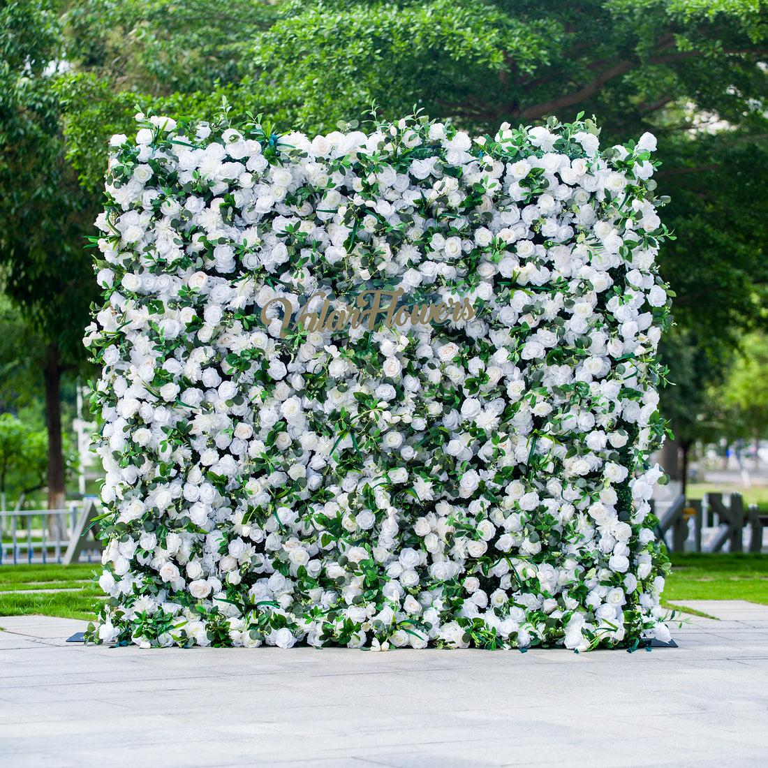 This photo showcases a large, square floral backdrop densely covered with white flowers, likely roses, interspersed with lush green foliage. The text “ValarFlowers” in elegant golden lettering stands out prominently. Ideal for events like a “Pure Elegance” wedding or a formal garden party, it exudes a sophisticated and serene atmosphere, perfect for upscale outdoor celebrations.