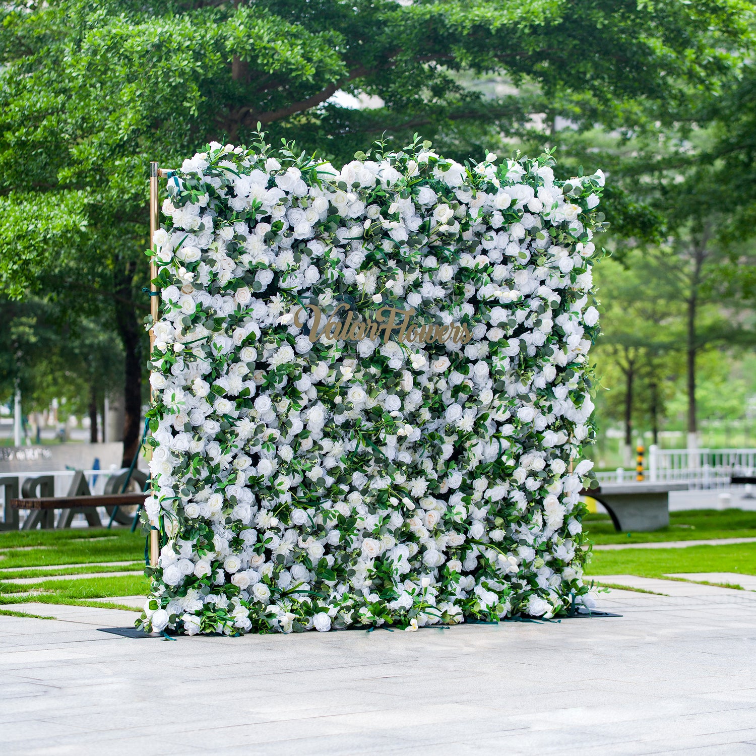 This photo features a large, square floral backdrop densely covered with white flowers, likely roses, interspersed with lush green foliage. The text “ValarFlowers” in elegant golden lettering stands out prominently. Ideal for events like a “White - Tie Garden Soirée” or a classic wedding, it exudes a sense of refined elegance, perfect for sophisticated outdoor celebrations.