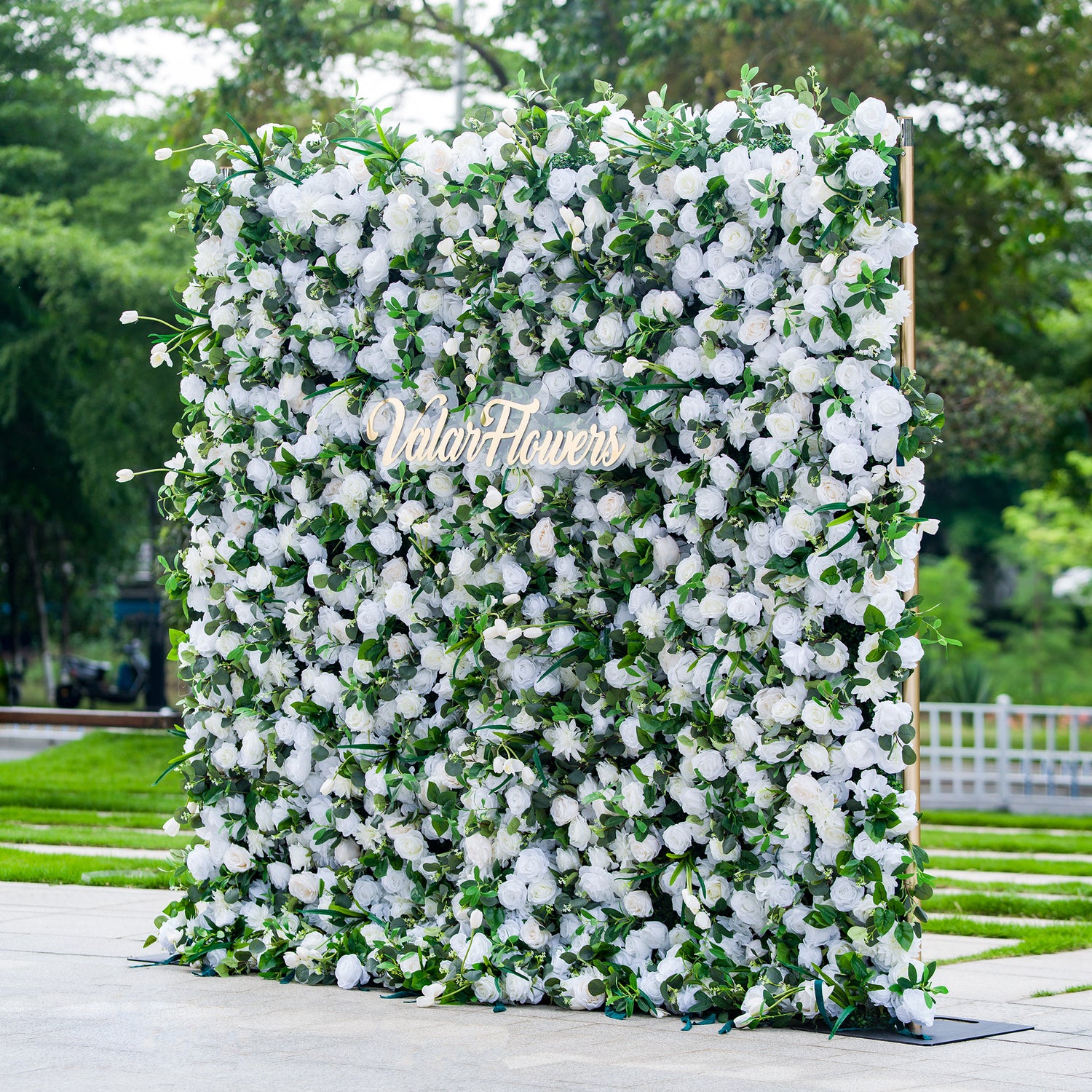 This photo showcases a large, square floral backdrop densely adorned with white flowers, likely roses and hydrangeas, interspersed with lush green foliage. The text “ValarFlowers” in elegant golden lettering stands out prominently. Ideal for events like a “White Elegance” wedding or a formal garden reception, it exudes a sense of purity and sophistication, perfect for upscale outdoor celebrations.
