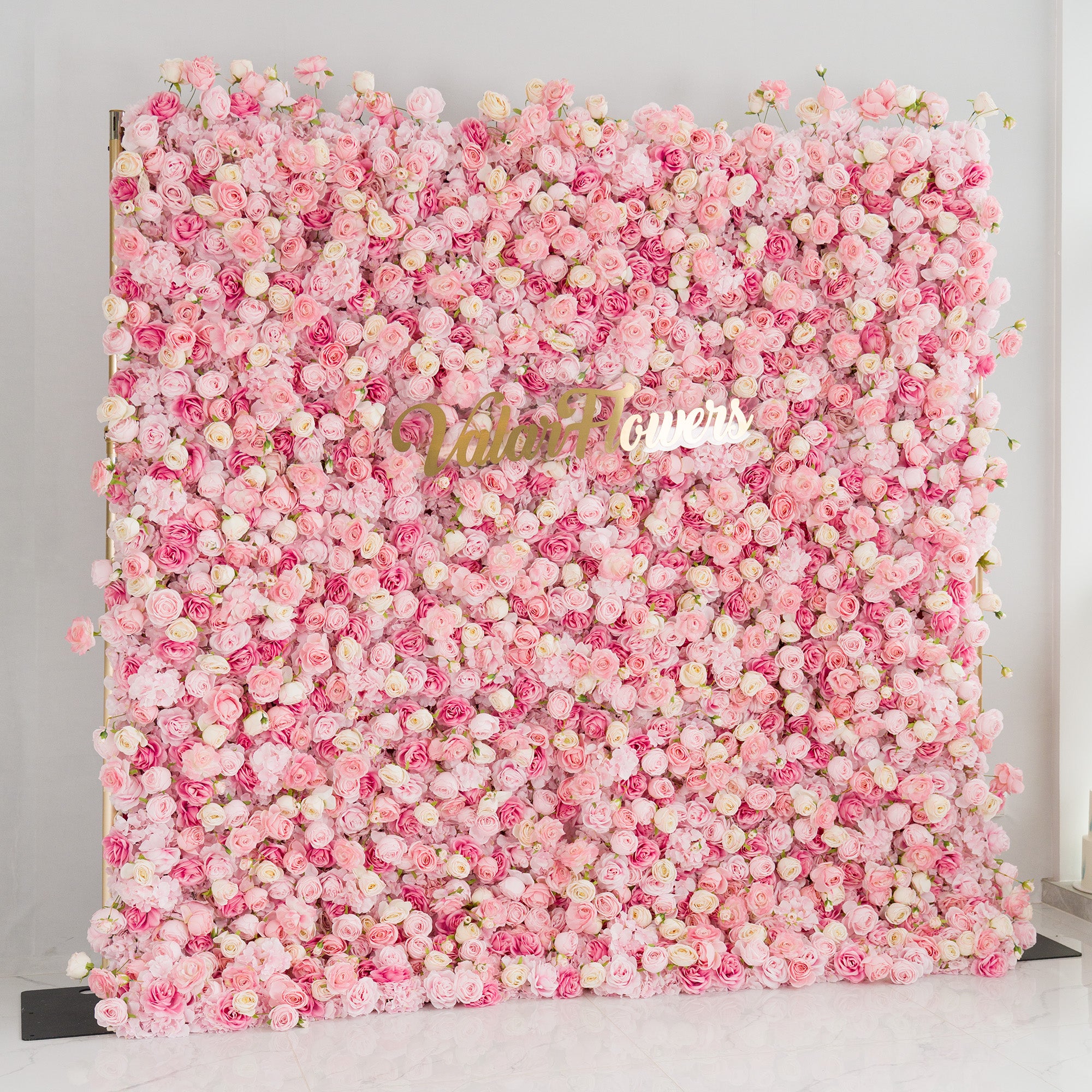 This photo showcases a large floral backdrop densely filled with roses in soft pink and white hues. The lush arrangement creates a dreamy, romantic atmosphere. Prominently displayed in the center is the “ValarFlowers” logo in elegant gold lettering. Ideal for weddings, bridal showers, or other romantic events, this backdrop exudes charm and sophistication, making it a stunning decorative piece.