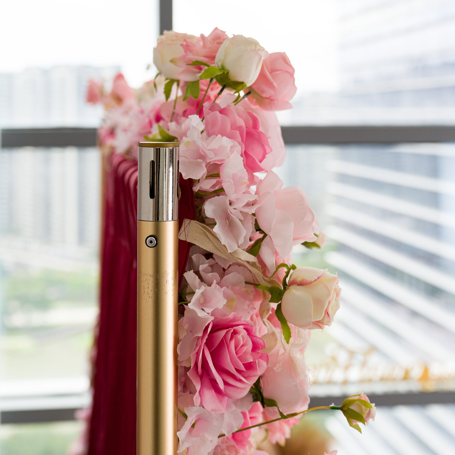 This photo captures a beautifully adorned floral arrangement. A golden pole is embellished with an assortment of pink and white roses, along with other delicate flowers. The blooms are lush and vibrant, set against a backdrop of a red fabric and a modern cityscape visible through a window. The combination of the golden pole, rich fabric, and fresh flowers creates an elegant and sophisticated scene, perfect for upscale events.