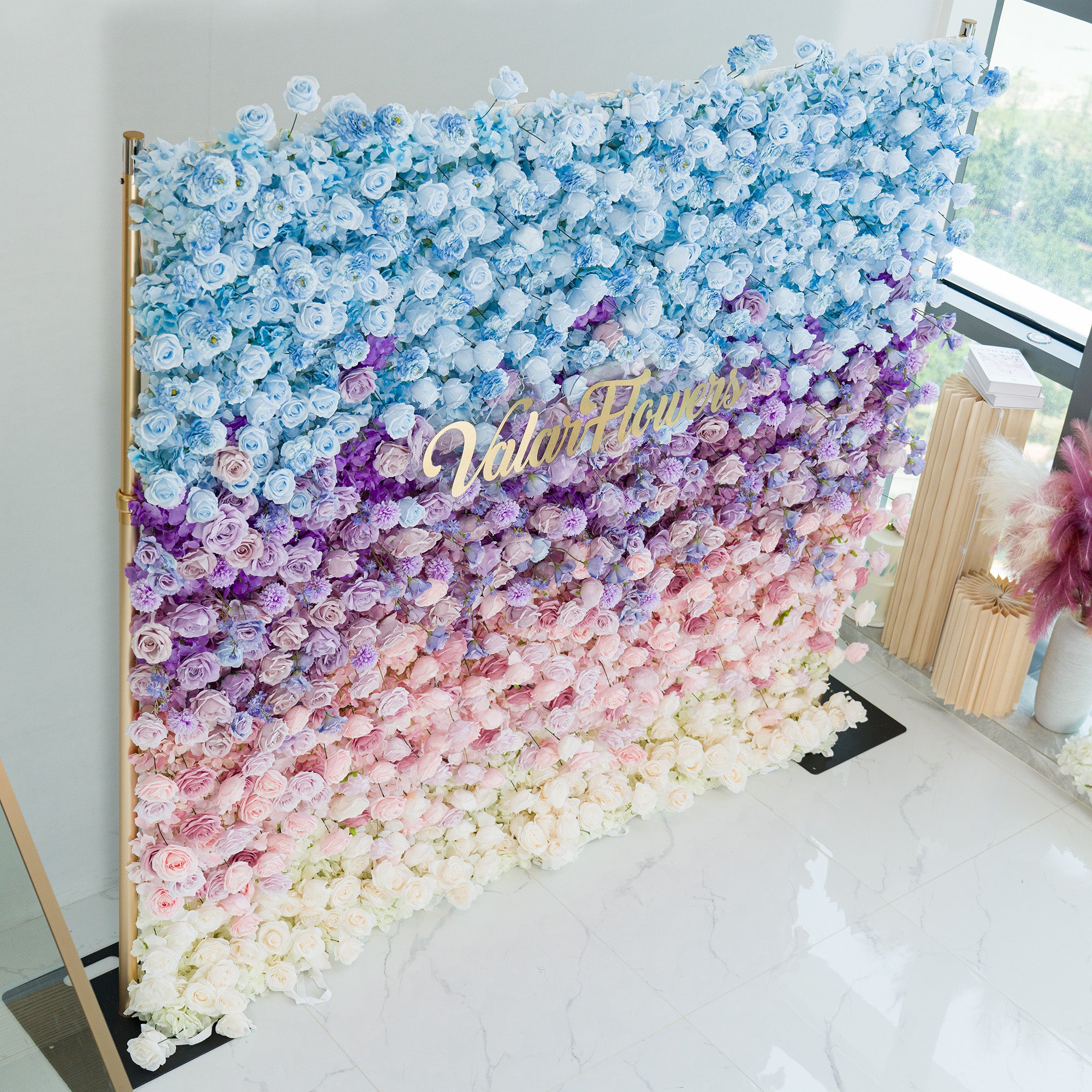 This elaborate floral wall showcases a gradient of roses and hydrangeas in hues of blue, purple, pink, and white. The ombre color theme, transitioning smoothly from sky - blue at the top to soft white at the base, creates a dreamy, romantic vibe. A gold “ValarFlowers” sign adds elegance. Perfect for weddings, galas, or brand events, it serves as a sophisticated, photo - worthy backdrop, blending natural beauty with polished design to elevate any occasion.