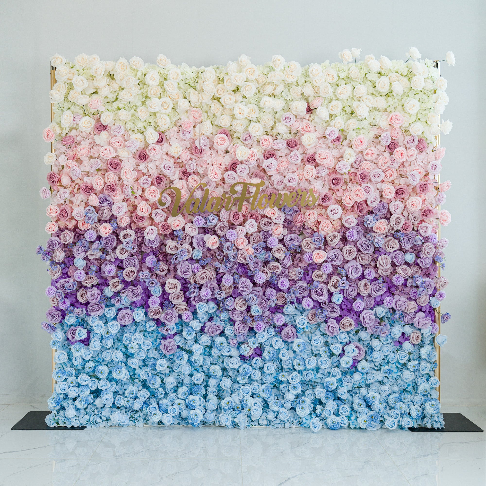 This stunning floral wall by ValarFlowers showcases a gradient of blooms—white roses, pink peonies, purple hydrangeas, and blue blossoms. The color theme, transitioning from white to blue through soft pastels, creates a dreamy, elegant vibe. Perfect for weddings, galas, or upscale events, it blends romance and sophistication, ideal as a photo backdrop or decorative centerpiece.