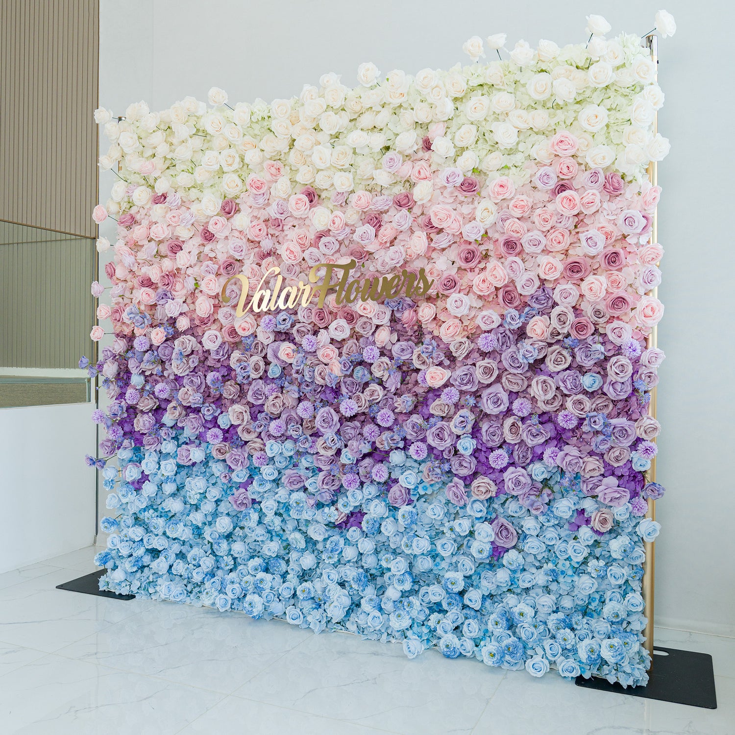 A stunning floral wall by ValarFlowers showcases a gradient of blooms—white roses at the top, transitioning through pink, purple, to blue flowers. The gold “ValarFlowers” logo adds elegance. Set against a minimalist backdrop, this lush, colorful display is perfect for events like weddings or photo shoots, blending romance and sophistication.