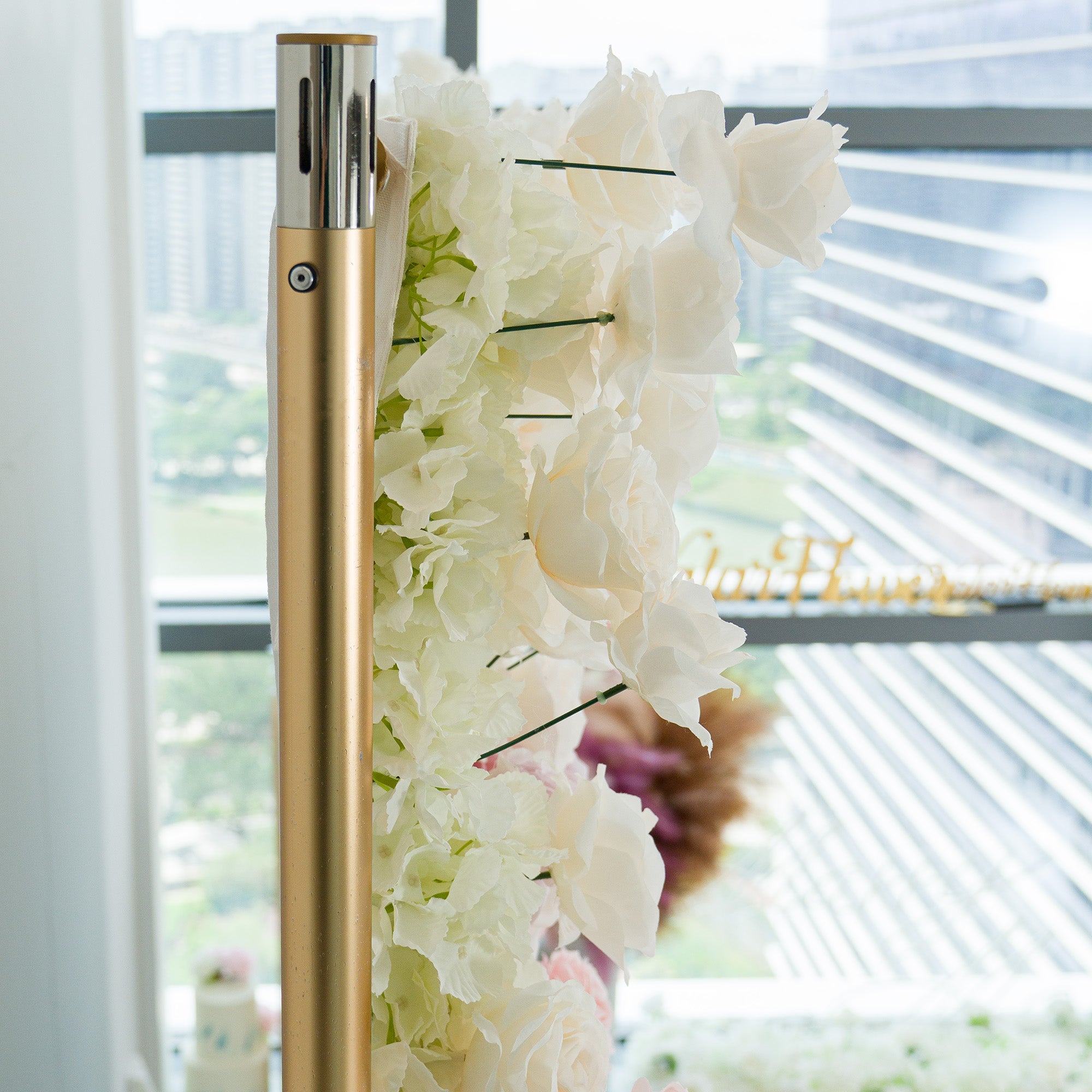 A gold - toned pole adorned with white hydrangeas and roses, paired with flowing fabric. Set against modern glass windows showing an urban backdrop, this elegant floral arrangement exudes a sophisticated, romantic vibe—ideal for weddings or upscale events, blending natural beauty with contemporary style.