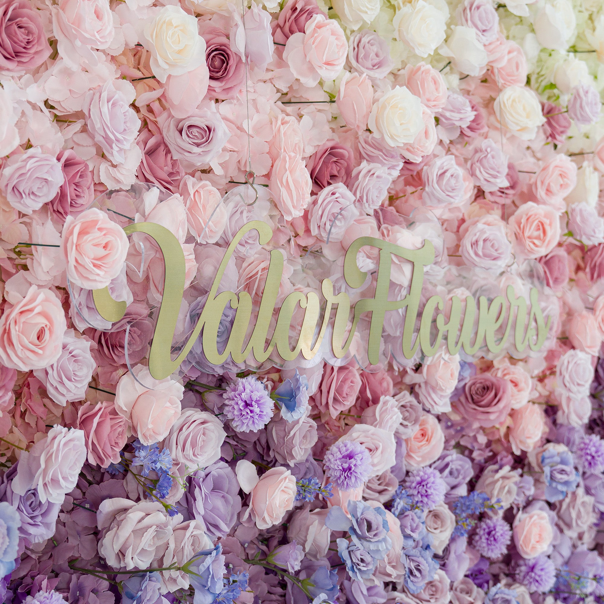 A lush floral wall with soft pink, lavender, and purple roses, plus delicate filler flowers. The gold “ValarFlowers” sign adds elegance. The pastel color theme creates a romantic, dreamy vibe—perfect for weddings, events, or brand activations, blending natural beauty with a polished aesthetic.