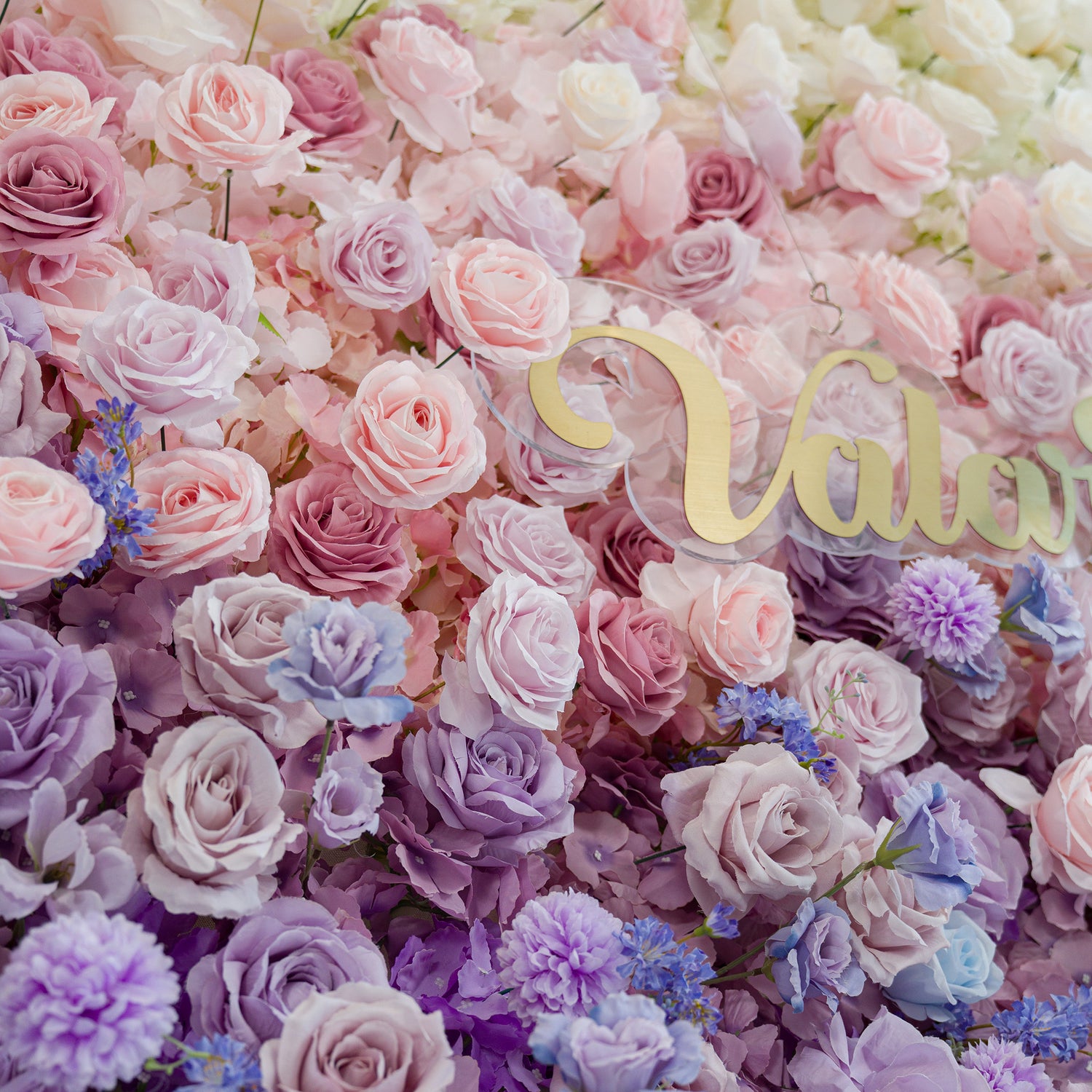 A lush floral wall bursts with pastel roses—soft pink, lavender, and purple—plus delicate blue blooms. The gold “Valar” sign adds elegance. The dreamy color theme creates a romantic, whimsical vibe, perfect for weddings, events, or photo backdrops, blending natural beauty with a polished touch.