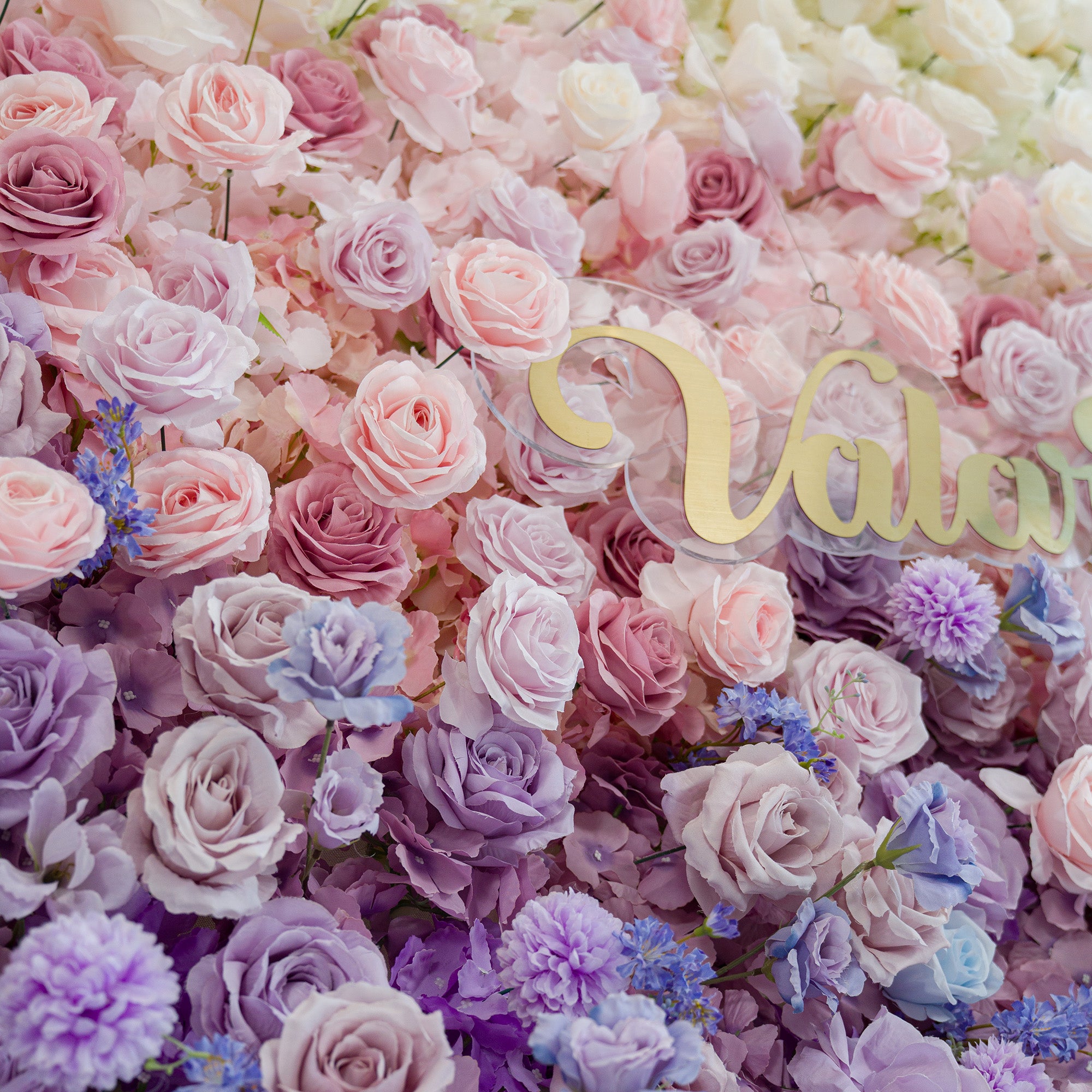 A lush floral wall bursts with pastel roses—soft pink, lavender, and purple—plus delicate blue blooms. The gold “Valar” sign adds elegance. The dreamy color theme creates a romantic, whimsical vibe, perfect for weddings, events, or photo backdrops, blending natural beauty with a polished touch.