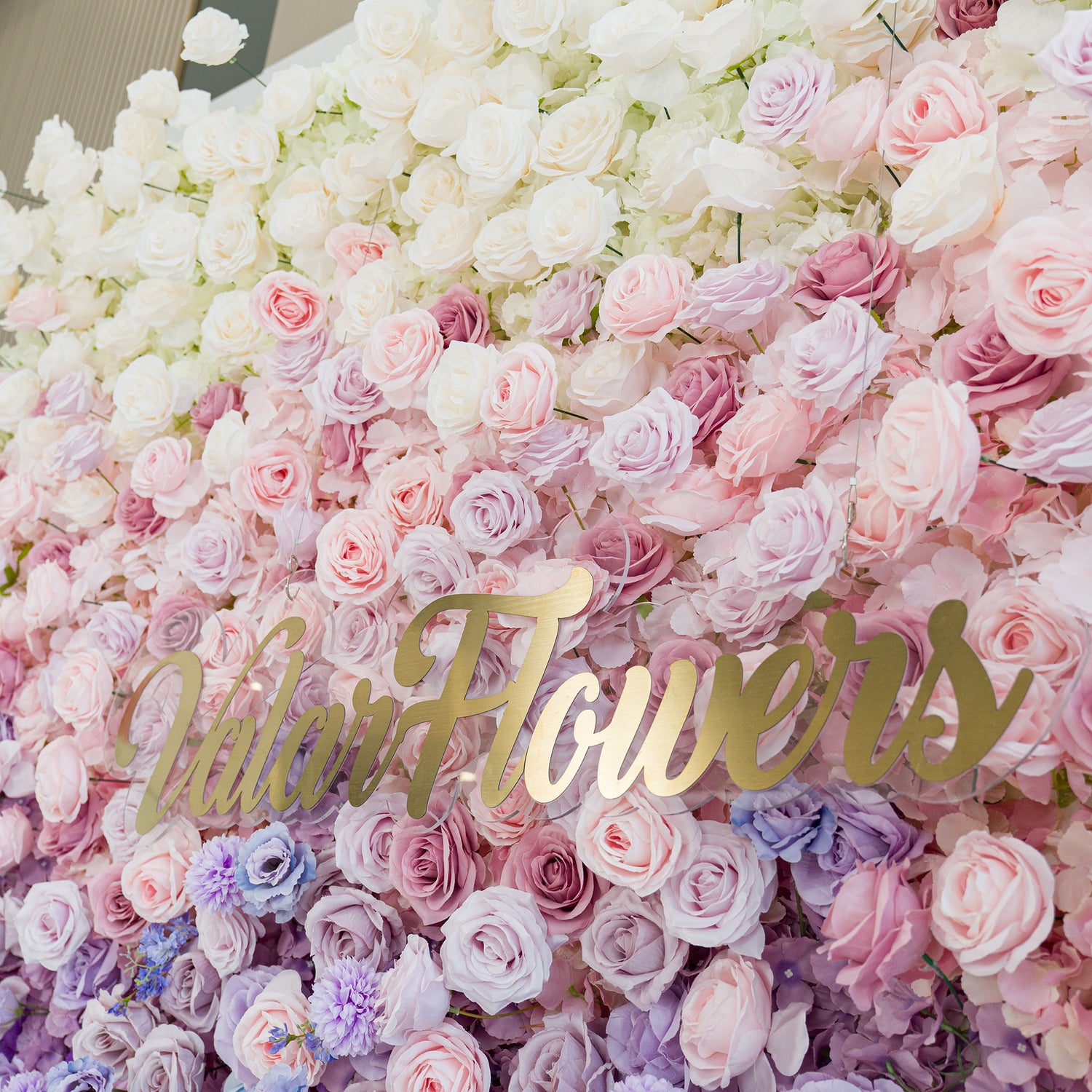 A lush floral wall features white roses, soft pink/lavender roses, and delicate filler flowers. The gold “ValarFlowers” sign adds elegance. The color theme—pastel gradients from white to purple—creates a romantic, dreamy vibe, perfect for weddings, events, or brand activations, blending natural beauty with polished charm.