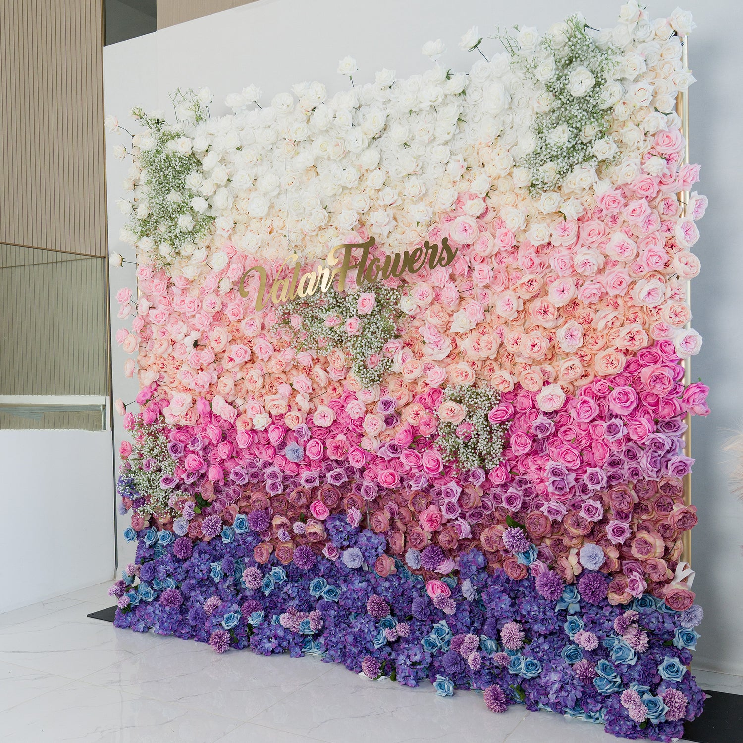 This stunning floral wall by ValarFlowers features a gradient of blooms—white roses and baby’s breath at the top, transitioning to soft pink, lavender, and deep purple flowers. The gold “ValarFlowers” logo adds elegance. Set indoors against a minimalist backdrop, it’s perfect for weddings, events, or photo backdrops, blending romance and sophistication.