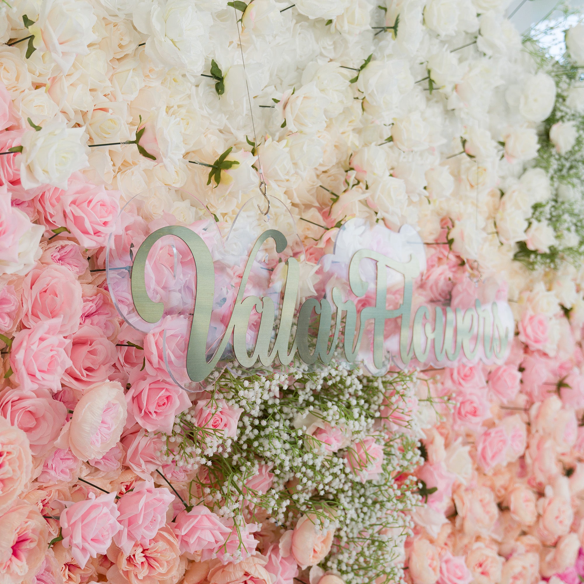This floral wall features white roses, soft pink roses, and baby’s breath. The color theme—white, blush pink, and delicate green—creates a romantic, dreamy vibe. Perfect for weddings, bridal showers, or elegant events, it blends classic blooms with a “ValarFlowers” acrylic sign, offering a timeless, sophisticated backdrop for photos and celebrations.