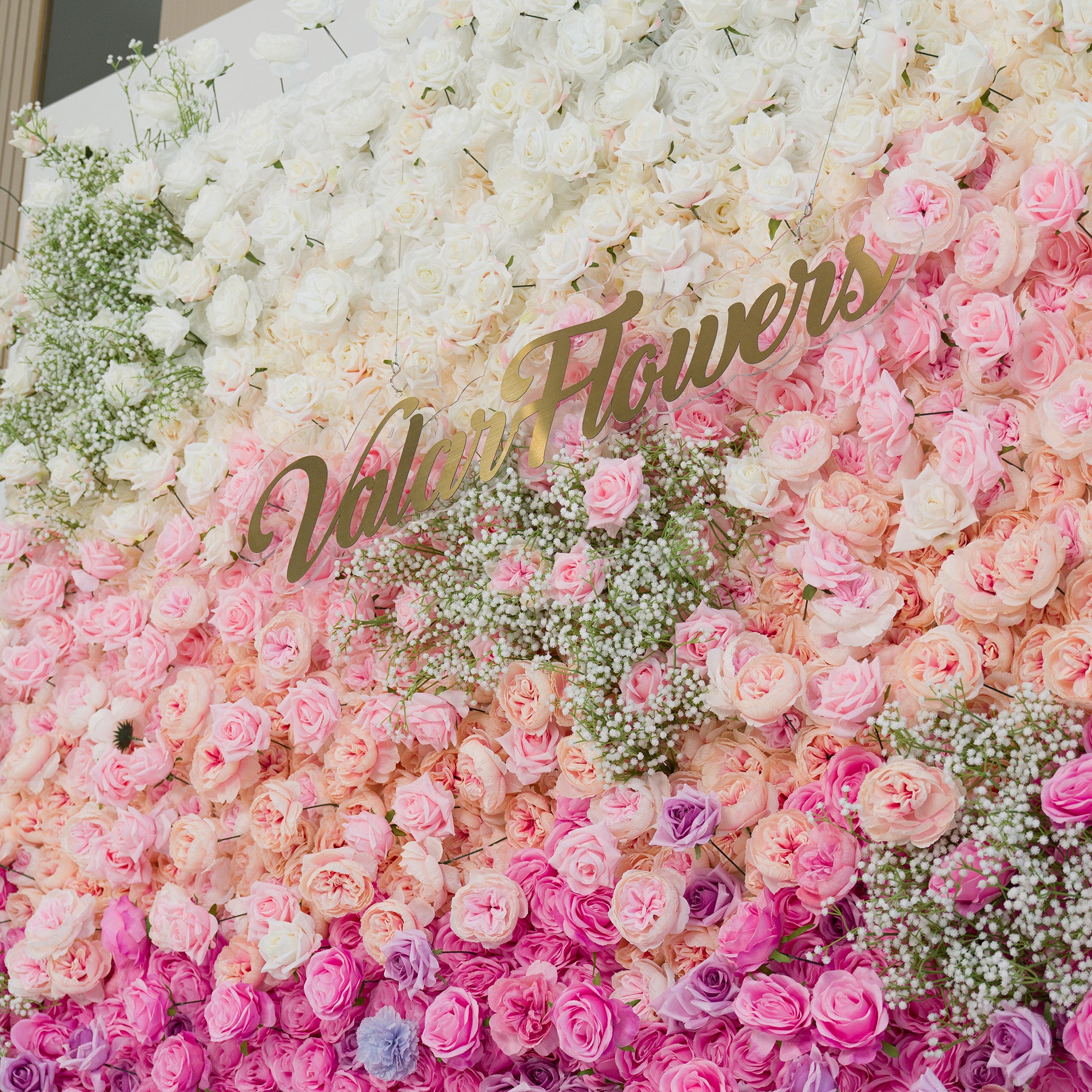 This stunning floral wall features white roses, baby’s breath, and gradient pink/purple blooms. The color theme—soft white to blush to deep pink/purple—creates a romantic, elegant vibe. Perfect for weddings, events, or brand activations, the “ValarFlowers” gold sign adds sophistication, blending natural beauty with a polished aesthetic.