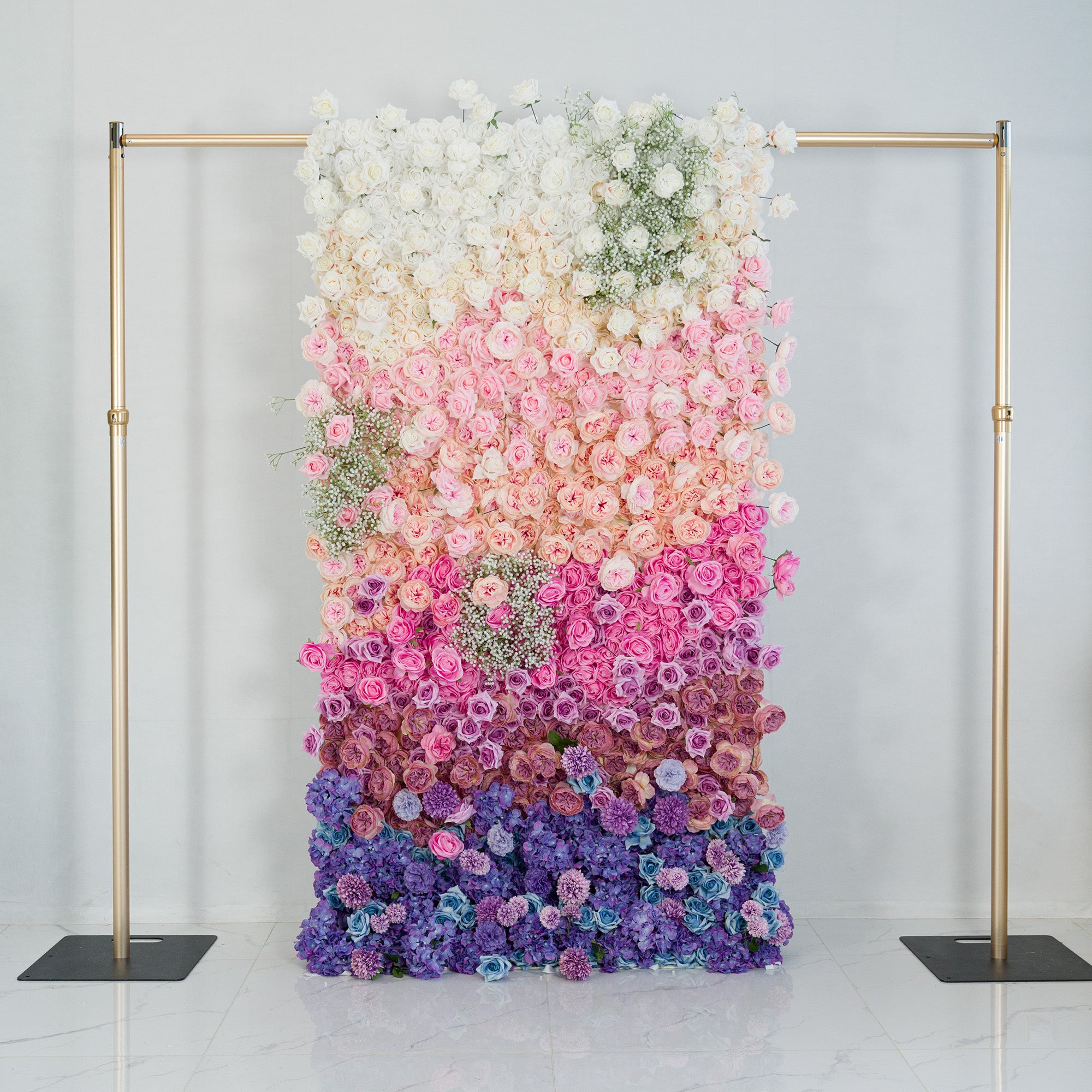 A 4x8ft vertical floral wall with a gradient of blooms—white roses at the top, transitioning to soft pink, lavender, and deep purple flowers, plus baby’s breath. Framed by gold poles, it’s set against a minimalist backdrop. Perfect for events like weddings or photo shoots, blending elegance and vibrant color in a sophisticated display.