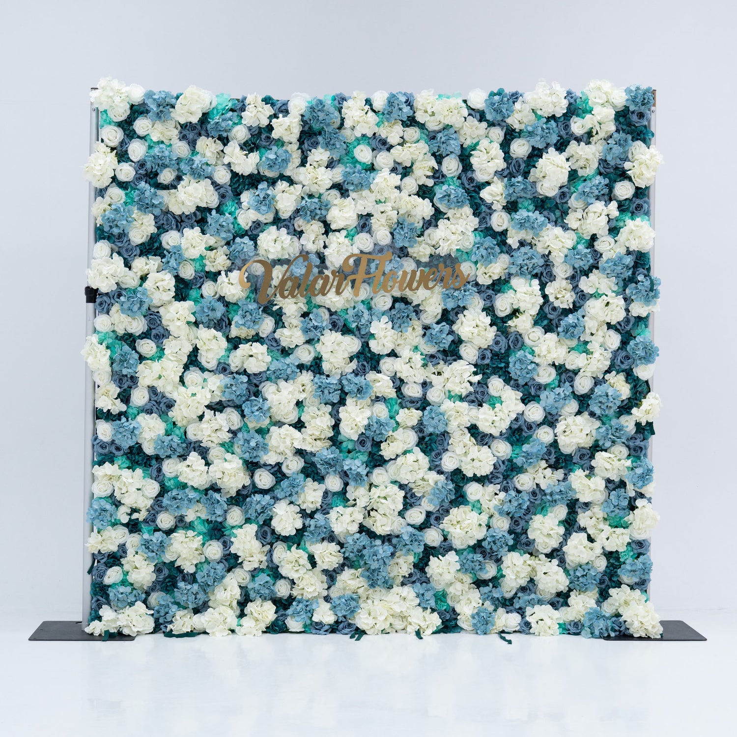 This photo showcases a stunning floral backdrop by Valar Flowers, featuring a dense arrangement of artificial roses in white and varying shades of blue. The backdrop is set against a plain white background, creating a clean and elegant look. The gold lettering of "Valar Flowers" adds a touch of sophistication, making it an ideal decorative piece for weddings, photo shoots, or other special events. The roses are meticulously arranged to create a visually appealing pattern, enhancing the overall aesthetic of
