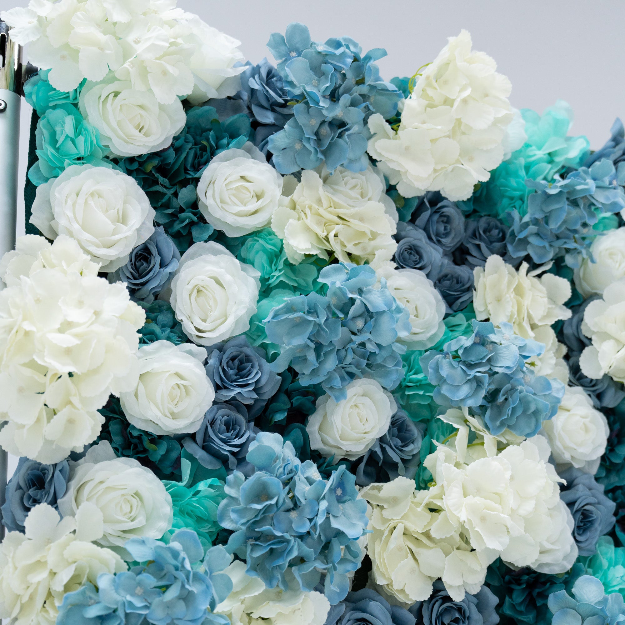 This photo showcases a lush arrangement of artificial flowers, featuring white and blue roses alongside blue hydrangeas. The flowers are densely packed, creating a visually appealing and vibrant display. The white roses stand out with their classic elegance, while the blue roses and hydrangeas add a cool, serene touch. This floral backdrop is perfect for weddings, photo shoots, or other special events, adding a touch of sophistication and natural beauty to any setting.