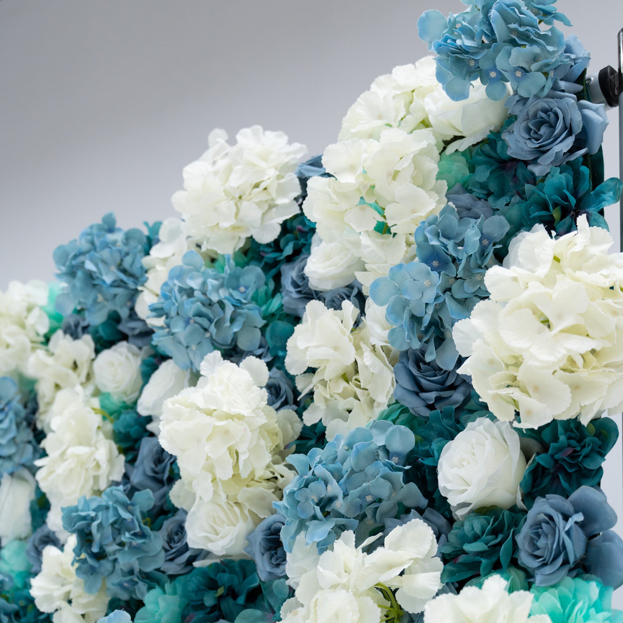 This photo showcases a lush arrangement of artificial flowers, featuring white and blue roses alongside blue hydrangeas. The flowers are densely packed, creating a visually appealing and vibrant display. The white roses stand out with their classic elegance, while the blue roses and hydrangeas add a cool, serene touch. This floral backdrop is perfect for weddings, photo shoots, or other special events, adding a touch of sophistication and natural beauty to any setting.