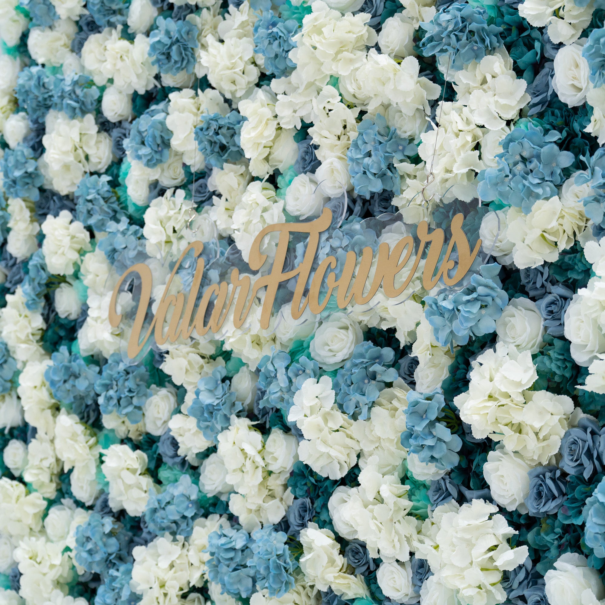 This photo features a stunning floral backdrop by Valar Flowers, showcasing a dense arrangement of artificial roses in white and varying shades of blue. The backdrop is set against a plain white background, creating a clean and elegant look. The gold lettering of "Valar Flowers" adds a touch of sophistication, making it an ideal decorative piece for weddings, photo shoots, or other special events. The roses are meticulously arranged to create a visually appealing pattern, enhancing the overall aesthetic of
