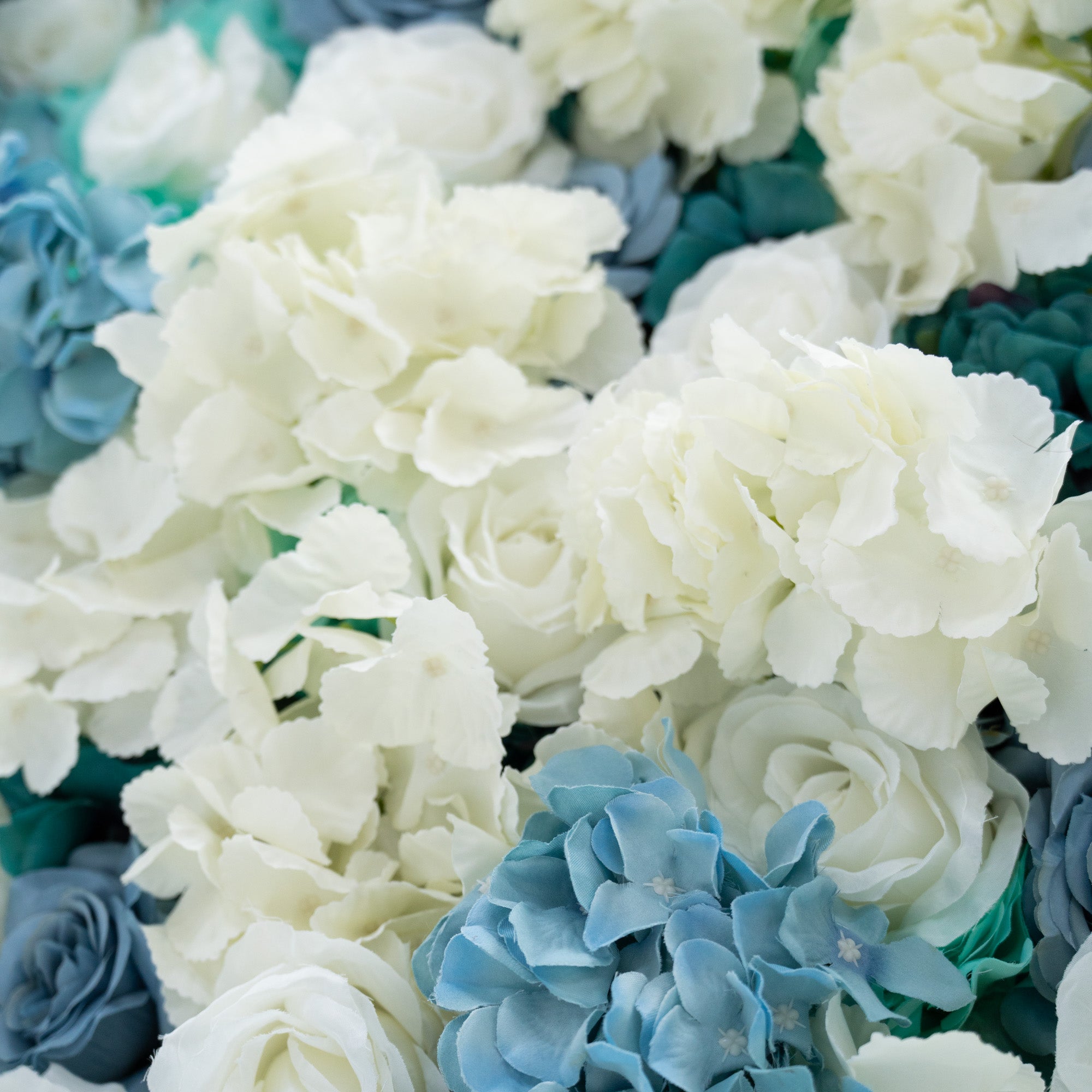 This photo presents a lush arrangement of artificial flowers, including white and blue roses as well as blue hydrangeas. The flowers are densely clustered, creating a visually attractive and vivid display. The white roses are elegant, while the blue roses and hydrangeas bring a calm and serene feel. This floral backdrop is perfect for weddings, photo shoots, or other special events with a blue and white color theme, adding a touch of sophistication and natural beauty to any setting.
