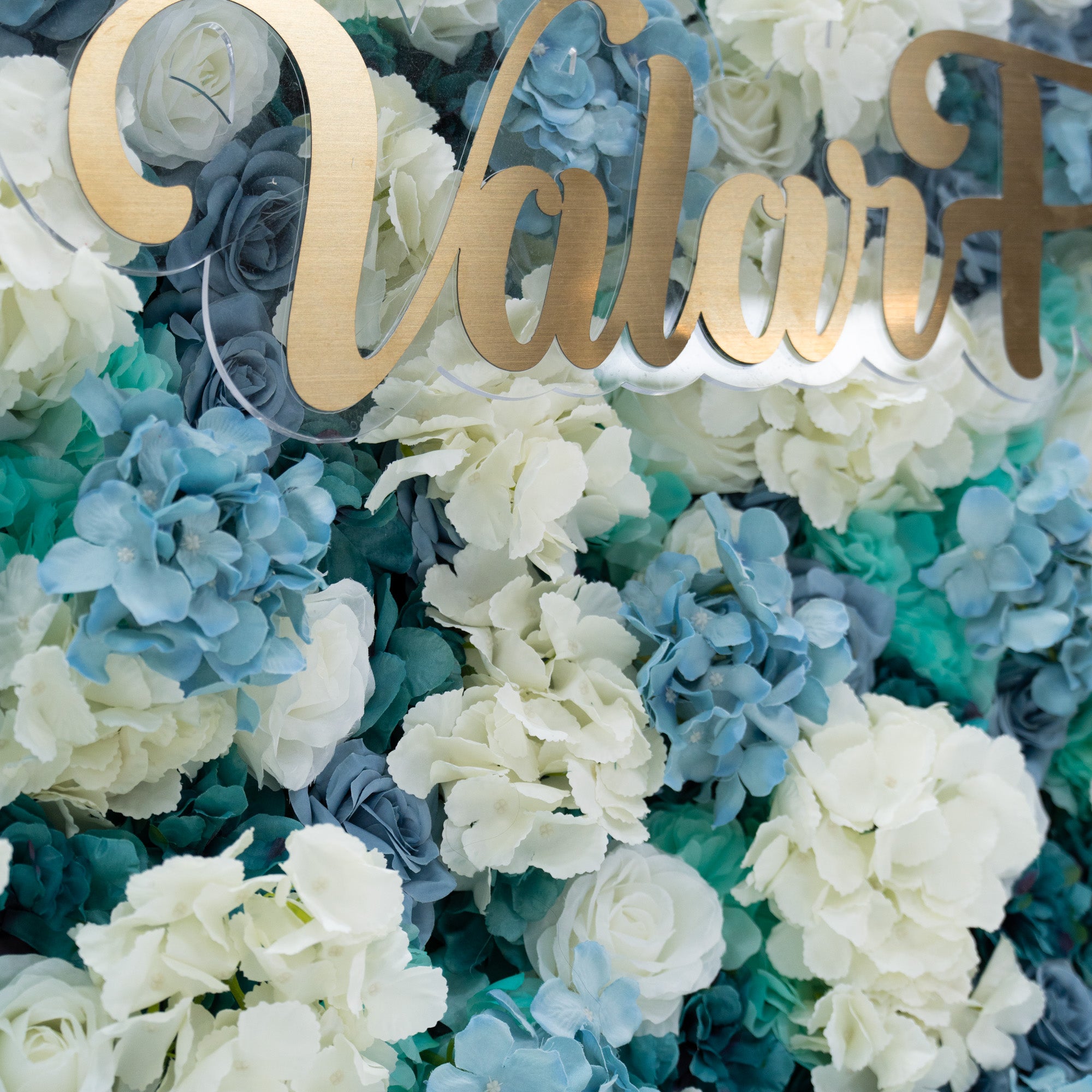 This photo showcases a lush arrangement of artificial flowers, featuring white and blue roses alongside blue hydrangeas. The flowers are densely packed, creating a visually appealing and vibrant display. The white roses stand out with their classic elegance, while the blue roses and hydrangeas add a cool, serene touch. This floral backdrop is perfect for weddings, photo shoots, or other special events with a blue and white color theme, adding a touch of sophistication and natural beauty to any setting.