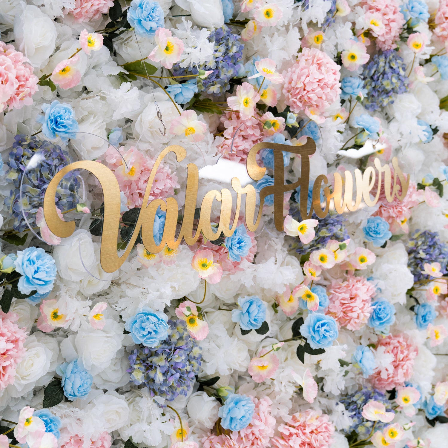 This photo features a stunning floral backdrop from Valar Flowers, showcasing a dense arrangement of artificial flowers in various shades of pink, blue, and white. The gold lettering of "ValarFlowers" adds a touch of elegance and sophistication, making it an ideal decorative piece for weddings, photo shoots, or other special events. The meticulous arrangement of the flowers enhances the overall aesthetic, adding a touch of natural beauty and charm to any setting.