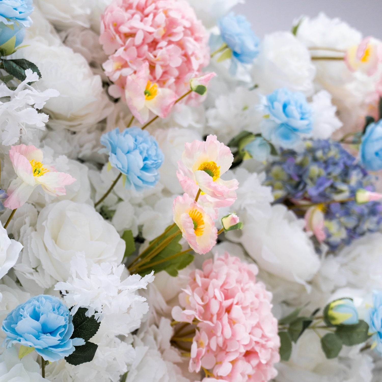 This photo showcases a beautiful arrangement of artificial flowers, featuring white and blue roses alongside pink and blue hydrangeas. The flowers are densely packed, creating a visually appealing and vibrant display. This floral backdrop is perfect for weddings, photo shoots, or other special events, adding a touch of sophistication and natural beauty to any setting.