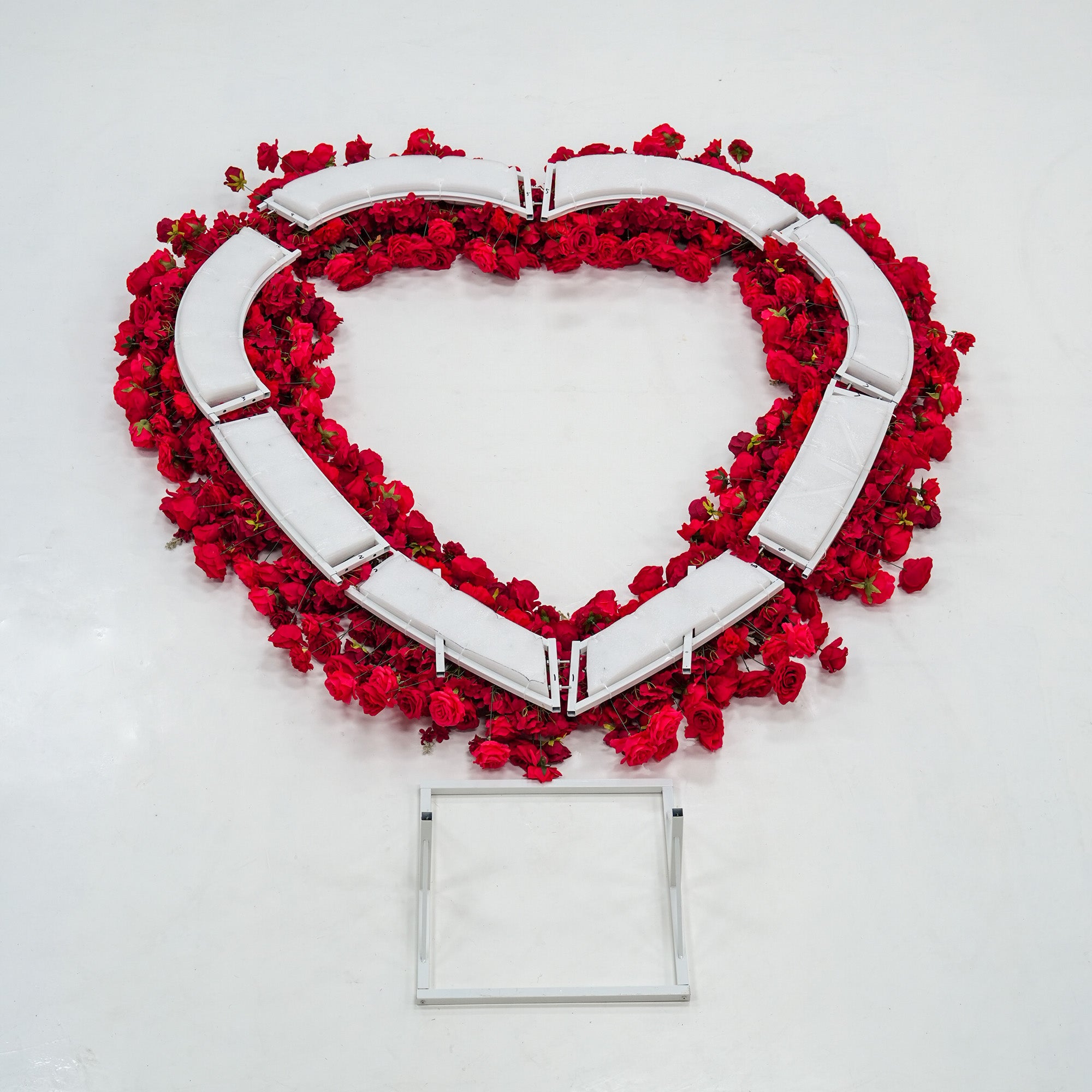 A heart - shaped structure with red roses adorning its edges, partially assembled with white frame pieces visible. Below it lies a square white metal frame. The vibrant red blooms contrast with the white components, suggesting a decorative floral installation in progress.