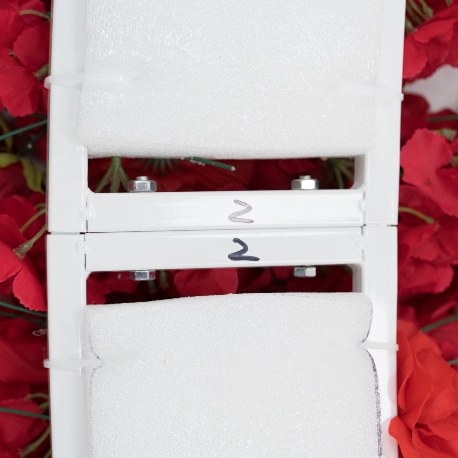 Close - up of two stacked white metal frames, each labeled “2” and padded with foam. Surrounding them are red artificial flowers. The frames, with visible bolts, seem to be parts of a decorative floral structure, likely for assembly into a larger decorative piece.