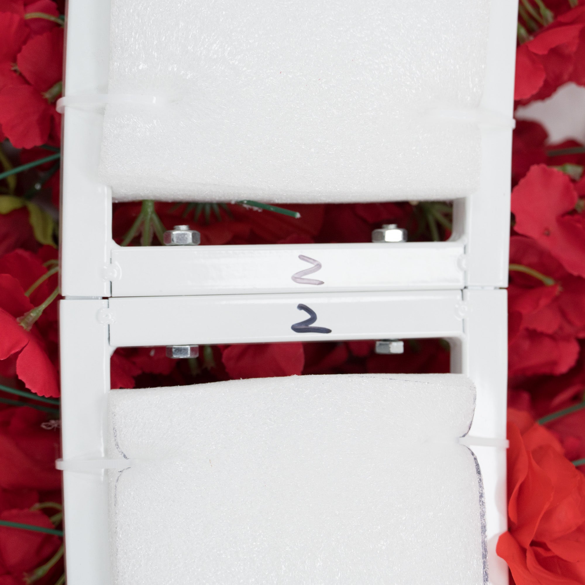 Close - up of two stacked white metal frames, each labeled “2” and padded with foam. Surrounding them are red artificial flowers. The frames, with visible bolts, seem to be parts of a decorative floral structure, likely for assembly into a larger decorative piece.