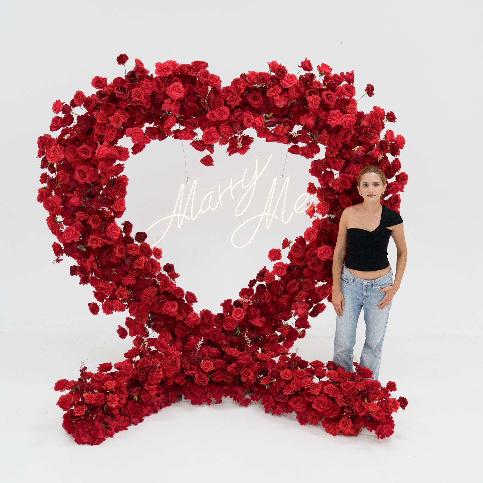 A woman in a black one - shoulder top and blue jeans stands beside a huge heart - shaped red rose installation. At its center, a glowing “Marry Me” sign adds a romantic touch. The lush, vibrant blooms create a striking, proposal - worthy scene.