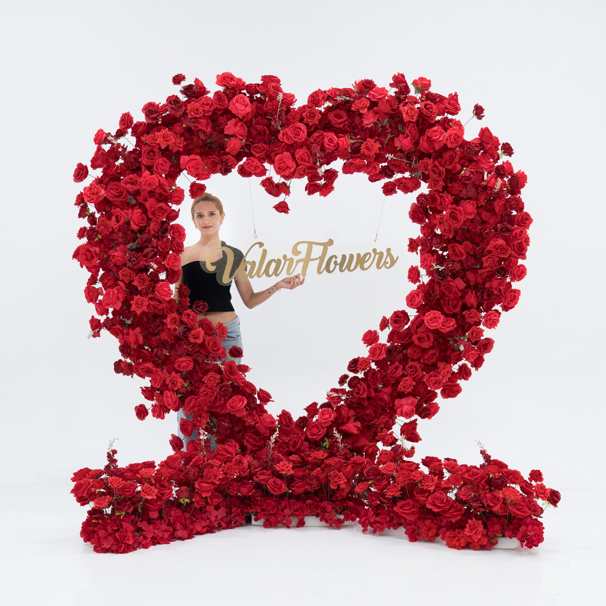 A woman stands within a large heart - shaped red rose installation. At its center, a gold “Valar Flowers” sign hangs. She wears a black one - shoulder top and blue jeans, posing with one hand extended. The lush, vibrant blooms create an eye - catching, decorative scene.