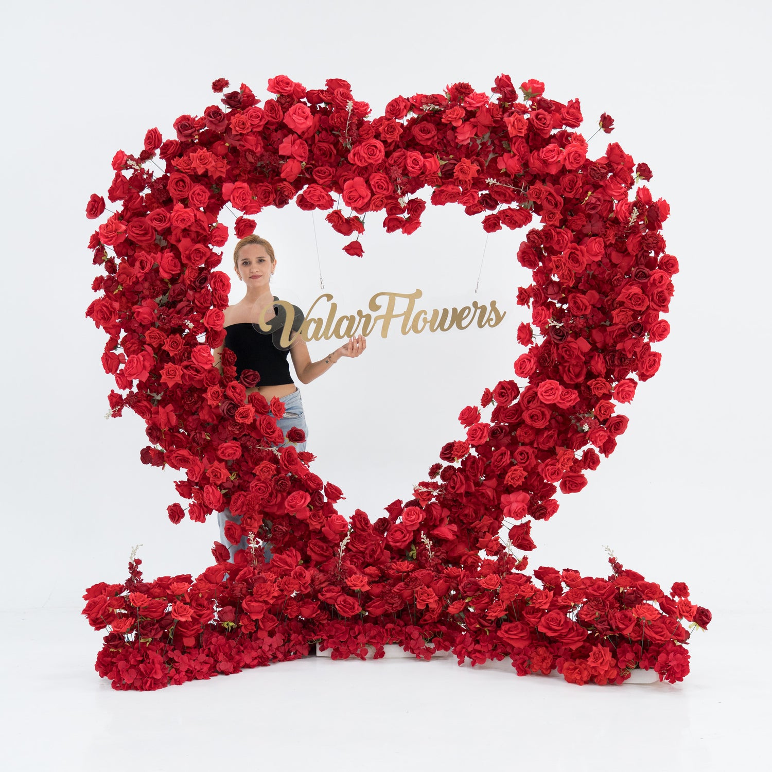 A woman stands inside a huge heart - shaped red rose installation. A glowing “Marry Me” sign hangs in the center. She wears a black one - shoulder top and blue jeans, with arms outstretched, creating a romantic and memorable proposal - style scene.