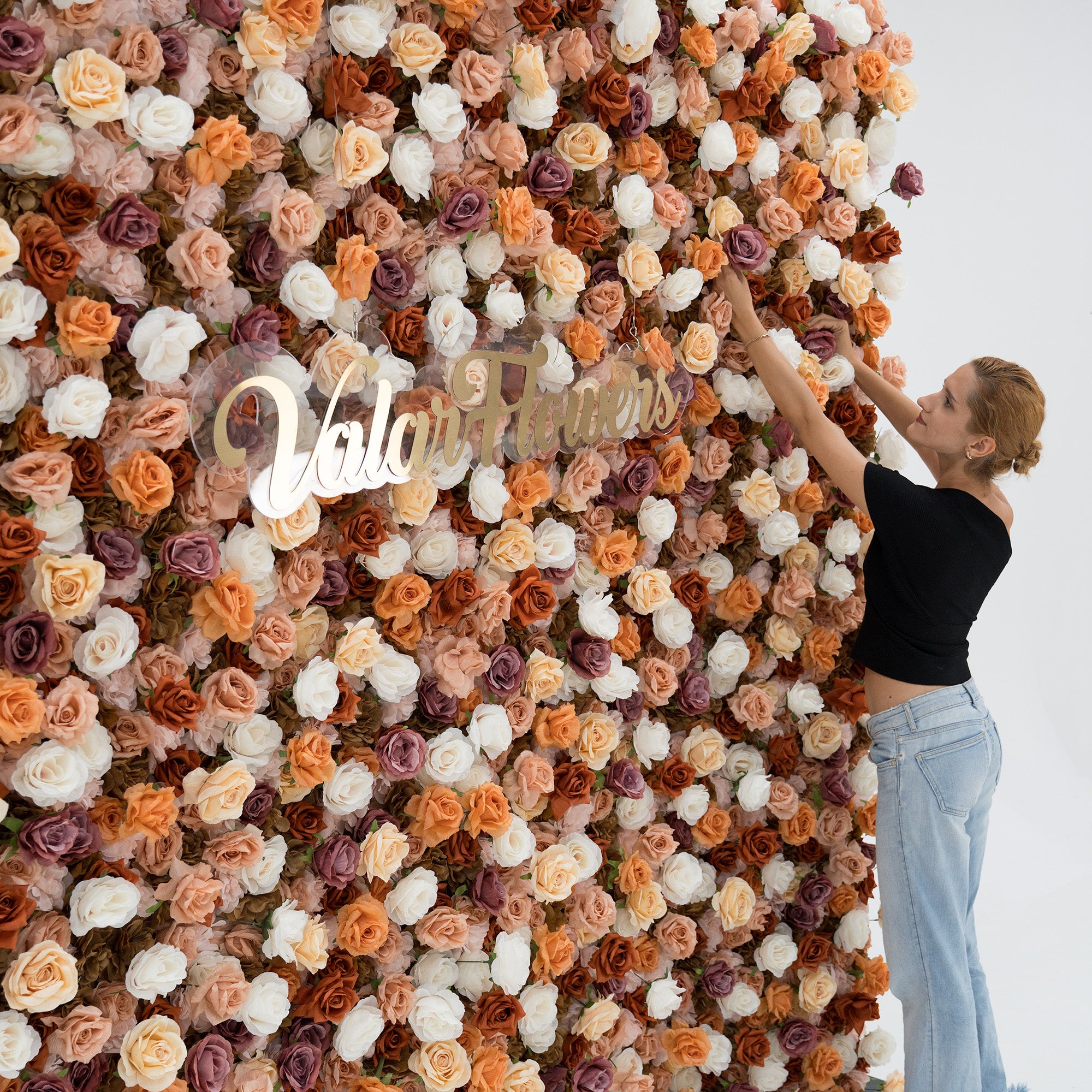 A woman in a black top and blue jeans adjusts a large floral backdrop. The backdrop features beige, white, and purple artificial flowers, with “ValarFlowers” in gold script. She adds a human touch to the elegant, textured display.