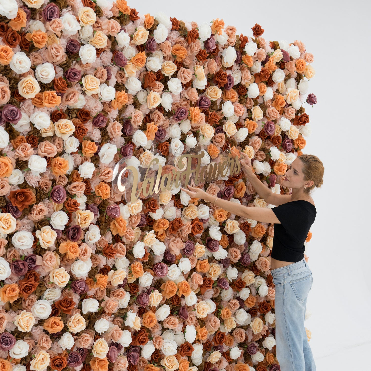 A woman in a black top and blue jeans adjusts a gold “ValarFlowers” sign on a lush artificial flower wall. The wall features a mix of beige, white, peach, and purple roses, creating an elegant, textured backdrop perfect for events or photo opportunities.