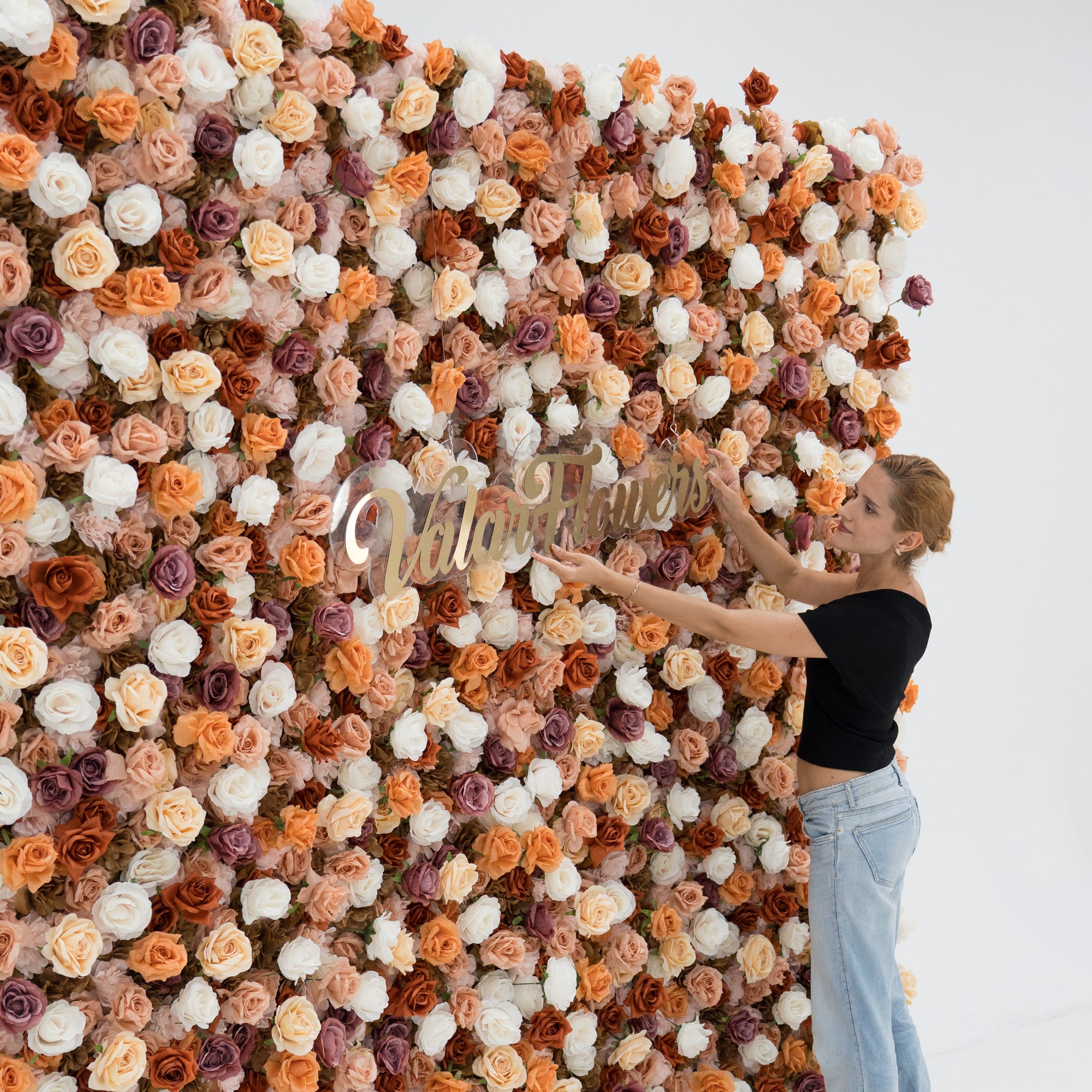 A woman in a black top and blue jeans adjusts a gold “ValarFlowers” sign on a lush artificial flower wall. The wall features a mix of beige, white, peach, and purple roses, creating an elegant, textured backdrop perfect for events or photo opportunities.