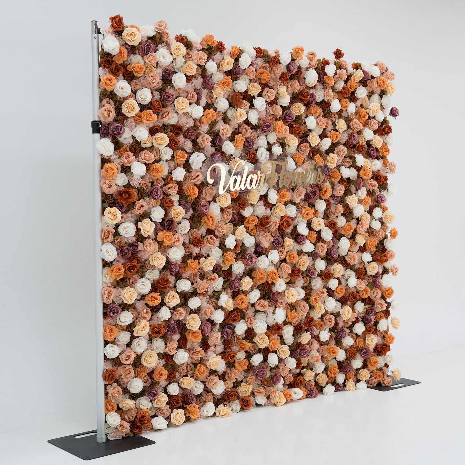 A large artificial flower wall features a mix of beige, white, and purple blooms. In the center, “ValarFlowers” is displayed in gold script. The wall is supported by metal poles and bases, creating an elegant, textured backdrop ideal for events or decor.