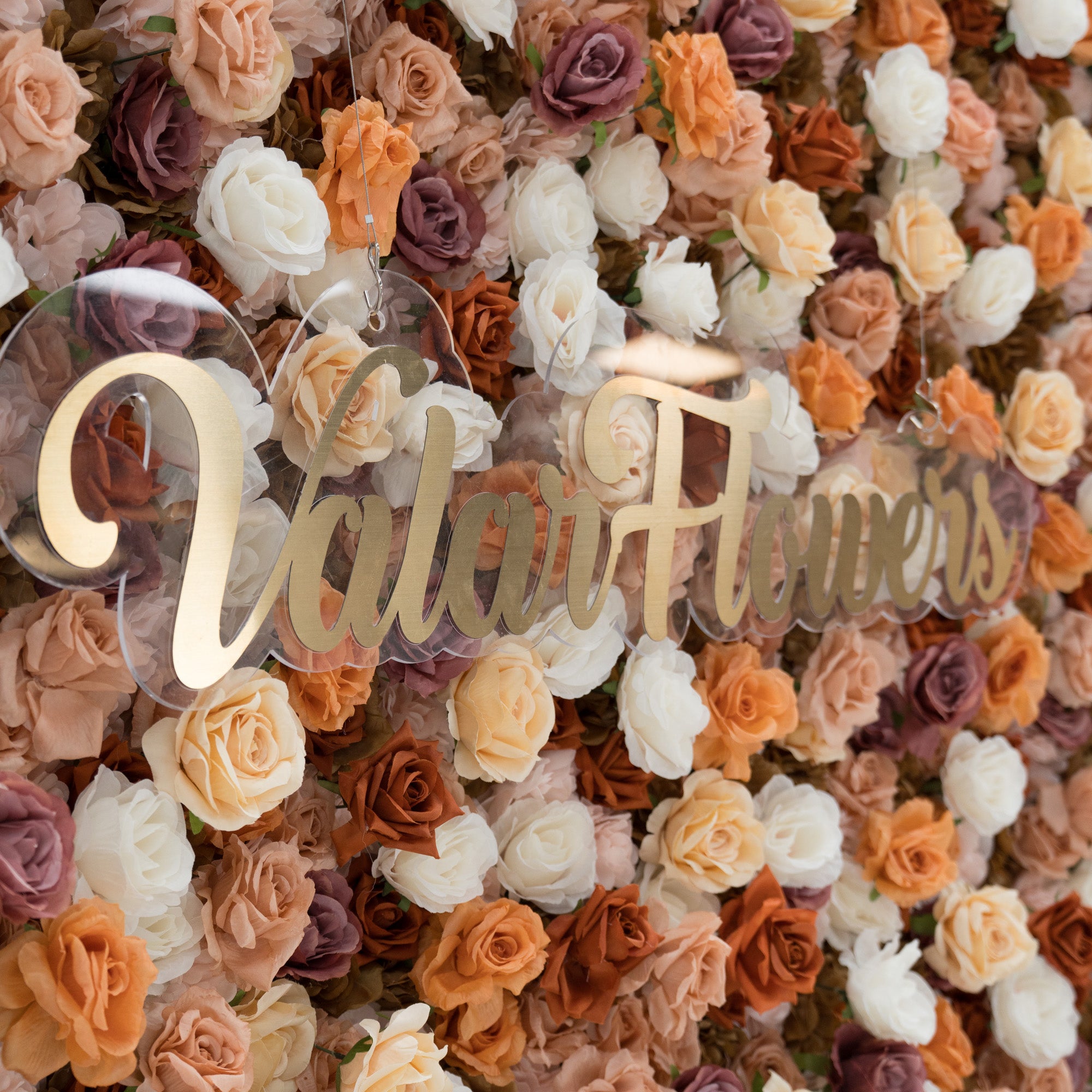 A close - up of a gold “ValarFlowers” sign against a lush artificial flower wall. The wall features beige, white, peach, and purple roses, creating a textured, elegant backdrop. The sign adds a branding element to the decorative floral display.