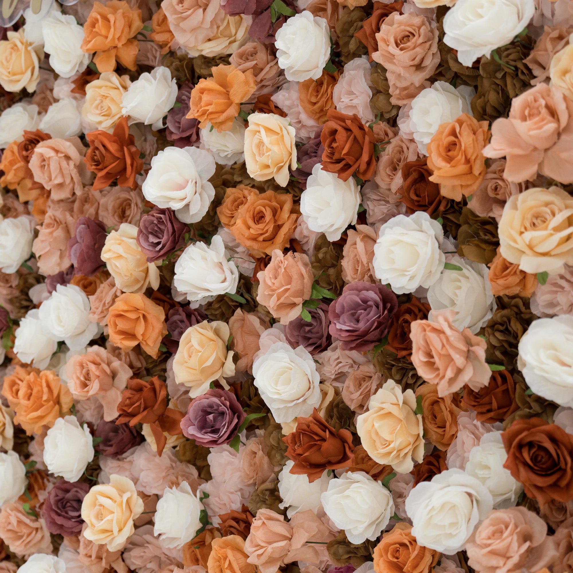 A close - up of a gold “ValarFlowers” sign against a lush artificial flower wall. The wall features beige, white, peach, and purple roses, creating a textured, elegant backdrop. The sign adds a branding element to the decorative floral display.
