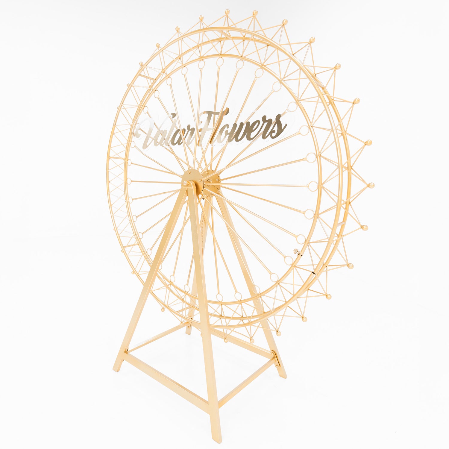 A golden metal Ferris wheel decoration showcases an open, intricate lattice design. With a triangular stand and circular spokes, it forms a delicate, ornamental structure. The shimmering gold finish exudes elegance, ideal for decorative event setups, and has “ValarFlowers” branding on it.