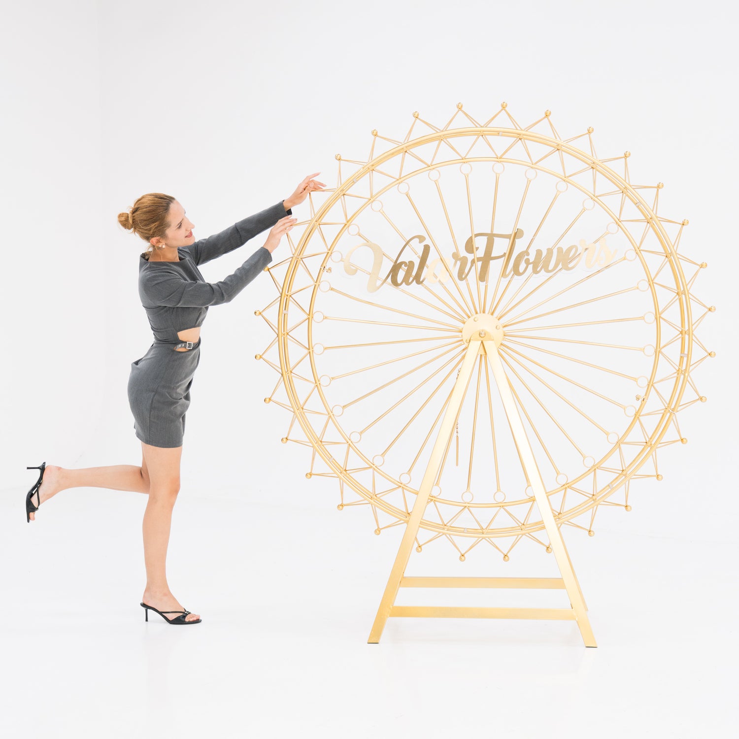 A woman in a gray, cut - out dress and black heels interacts with a large, golden Ferris wheel decoration. The wheel features intricate latticework and a “ValarFlowers” sign. The scene, set against a white backdrop, blends elegance and whimsy, ideal for event decor.