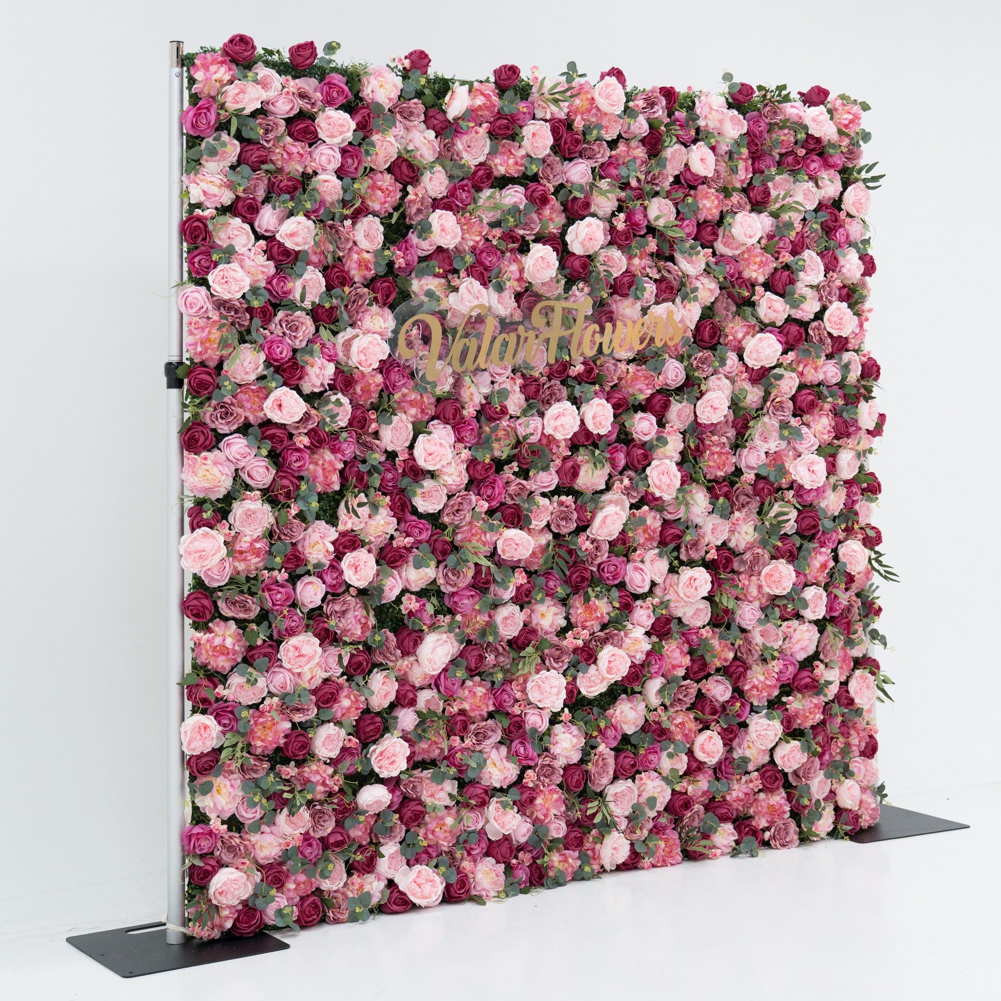 A large artificial flower wall, densely packed with pink and deep - pink roses, plus green foliage. A gold “ValarFlowers” sign is centrally placed, creating a romantic, elegant backdrop—ideal for events, exuding charm and sophistication with its lush, colorful blooms.