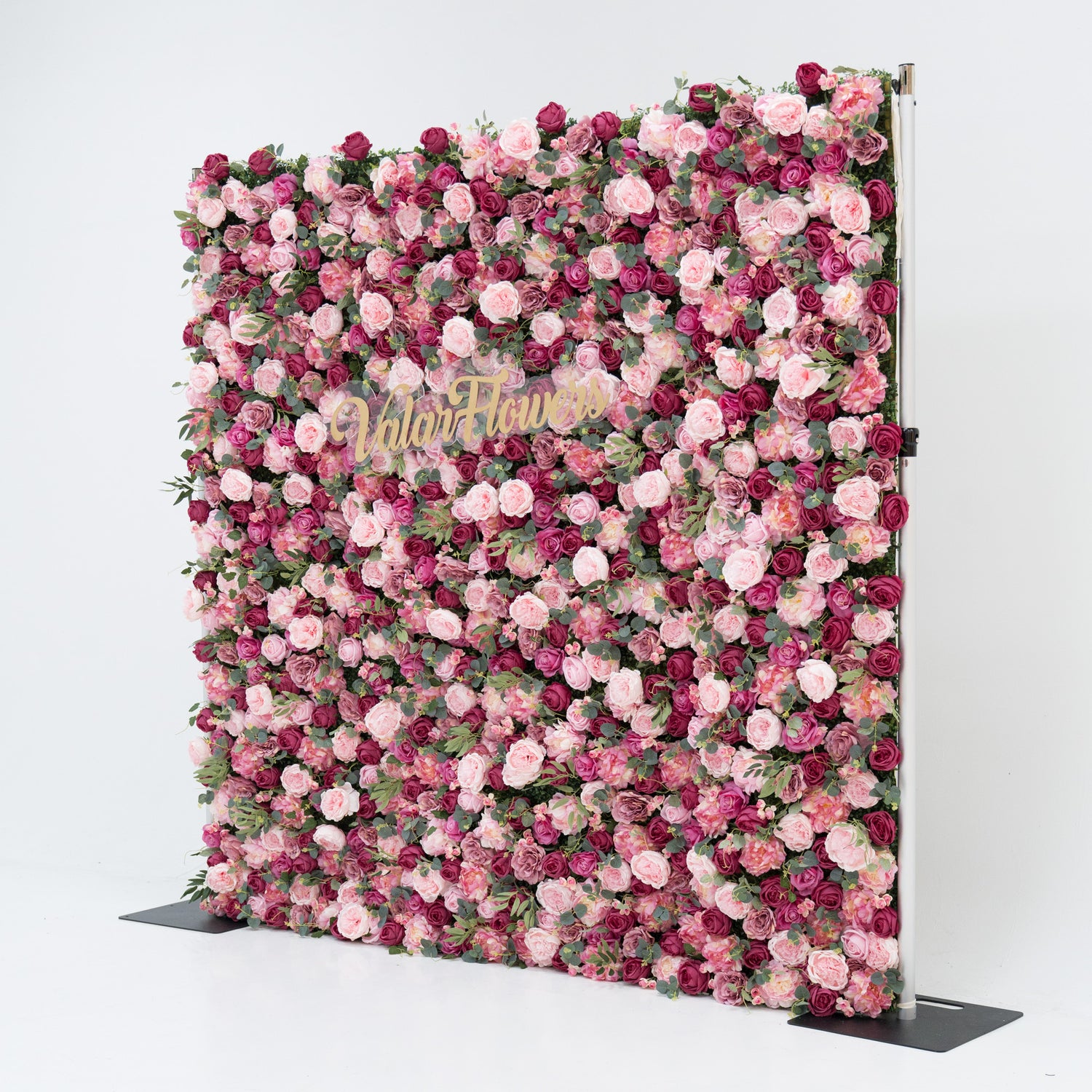 A large artificial flower wall, densely packed with pink and deep - pink roses, plus green foliage. A gold “ValarFlowers” sign is centered, creating an elegant, romantic backdrop—ideal for events, exuding charm and sophistication.
